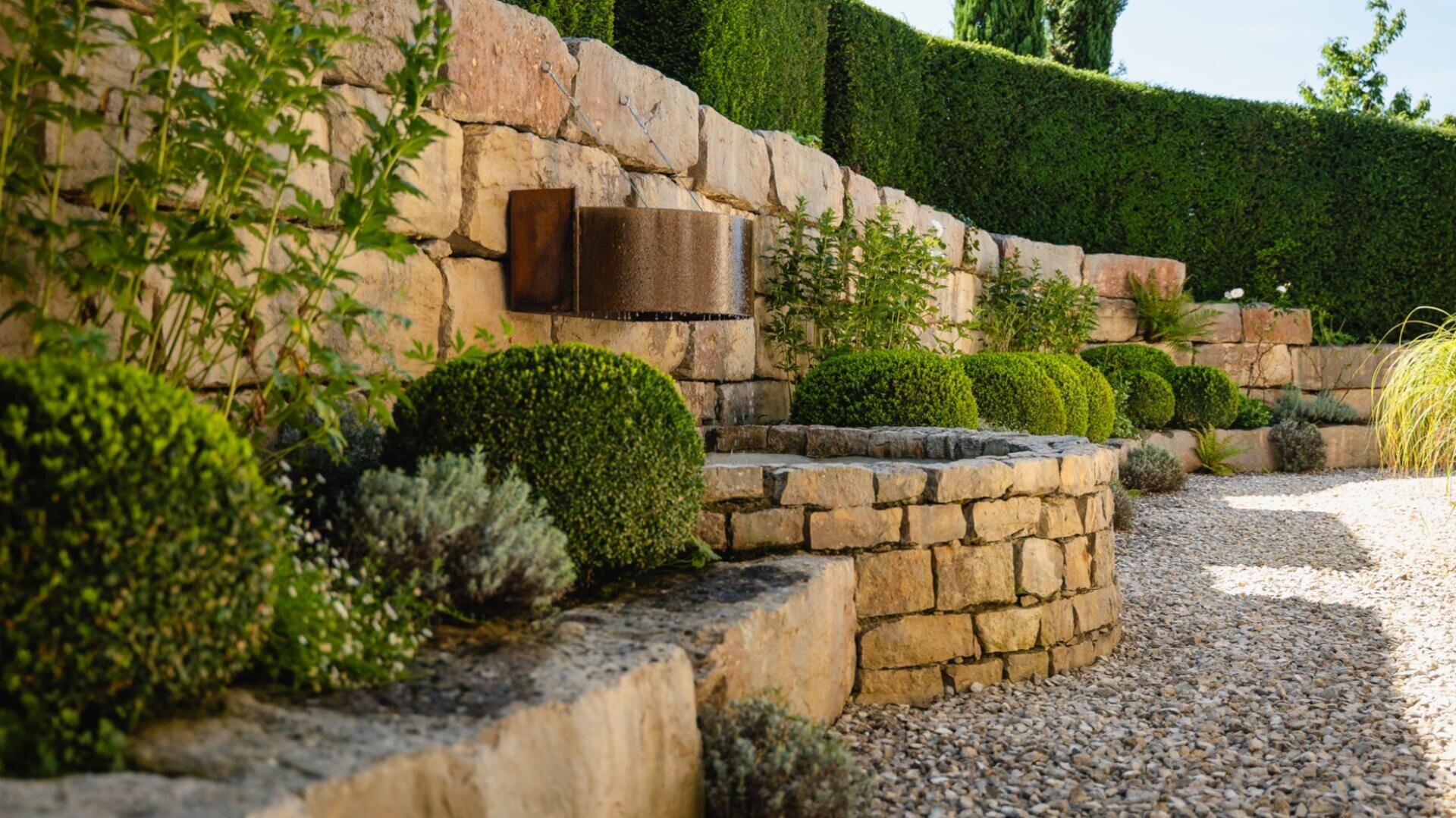 Ein terrassenförmig angelegter Garten mit steinernen Stützmauern, ordentlich gestutzten Rundsträuchern, kleinen Pflanzen und einem Schotterweg. Die Szenerie ist üppig und gepflegt, vor dem Hintergrund grüner Hecken und eines strahlend blauen Himmels.