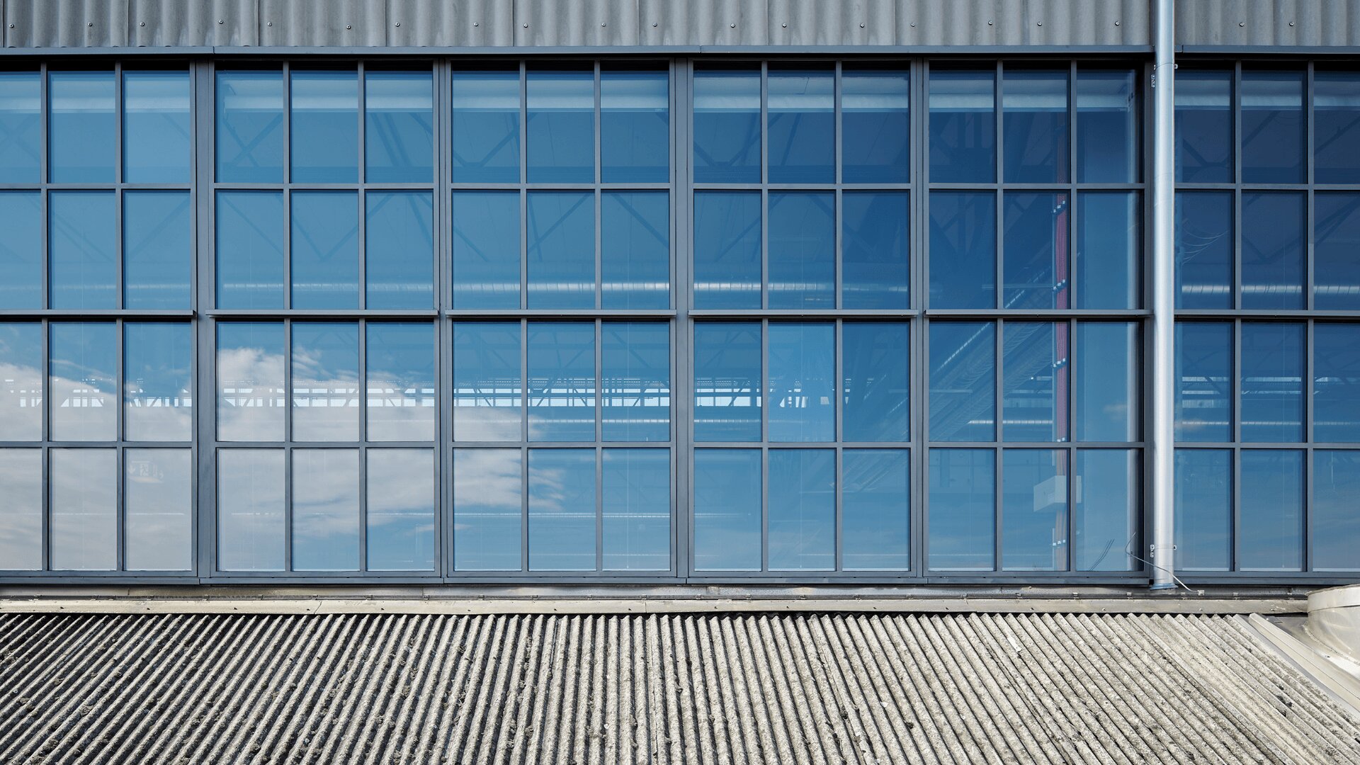Große Fenster im Industriestil mit blau getöntem Glas reflektieren den Himmel und die Wolken. Unter den Fenstern befindet sich ein Wellblechdach, und das Äußere des Gebäudes hat einen modernen, metallischen Look.