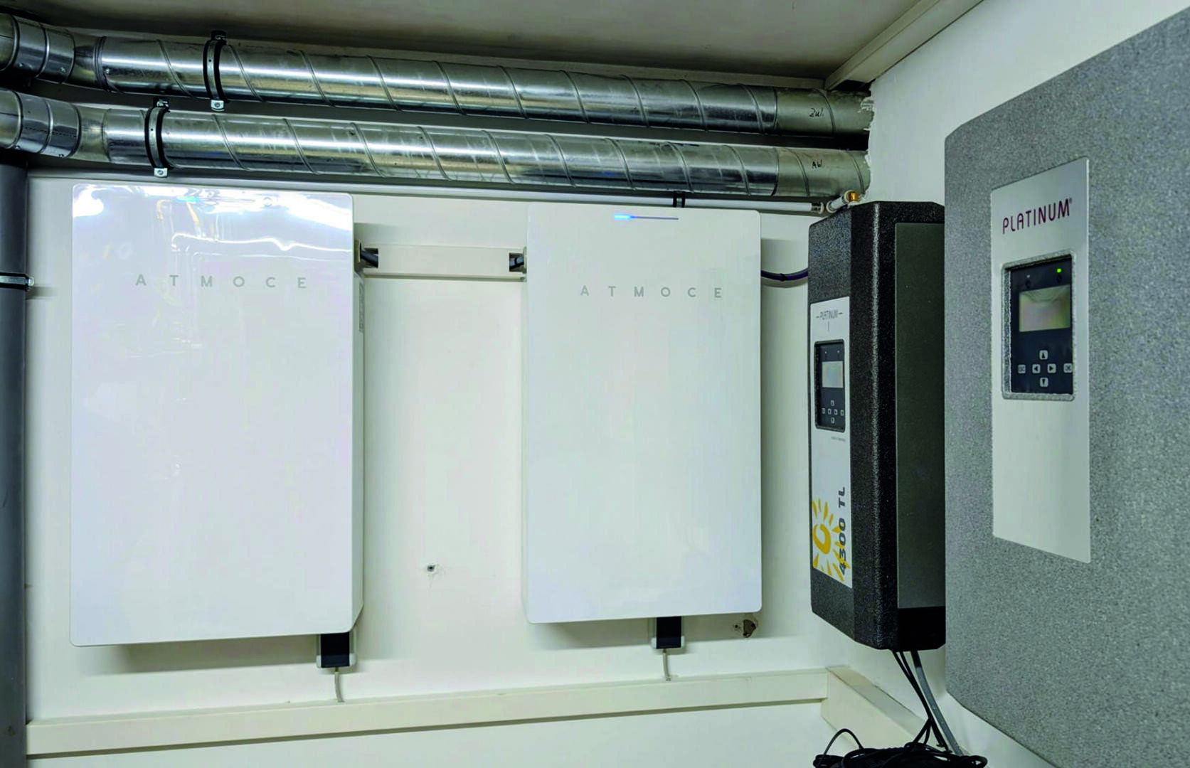 Zwei weiße, an der Wand montierte Geräte mit der Aufschrift ATMOCE sind nebeneinander installiert, darüber befinden sich Metallkanäle. Rechts daneben ist auf einer grauen Platte ein Bedienfeld mit der Aufschrift PLATINUM angebracht. Die Umgebung scheint ein Hauswirtschaftsraum zu sein.