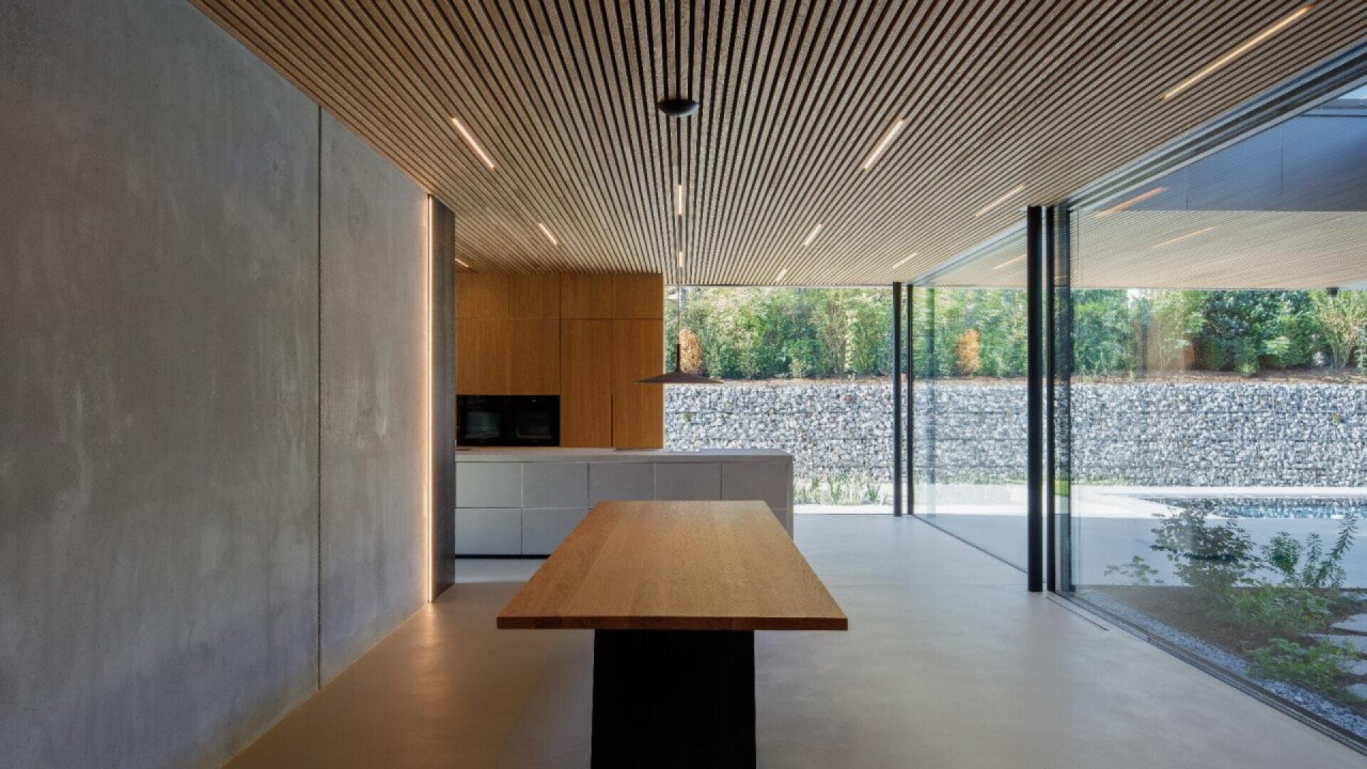 Cuisine ouverte moderne avec des murs en béton, des armoires en bois et un grand îlot en bois. De grandes fenêtres offrent une vue sur un mur de pierre et une végétation luxuriante.