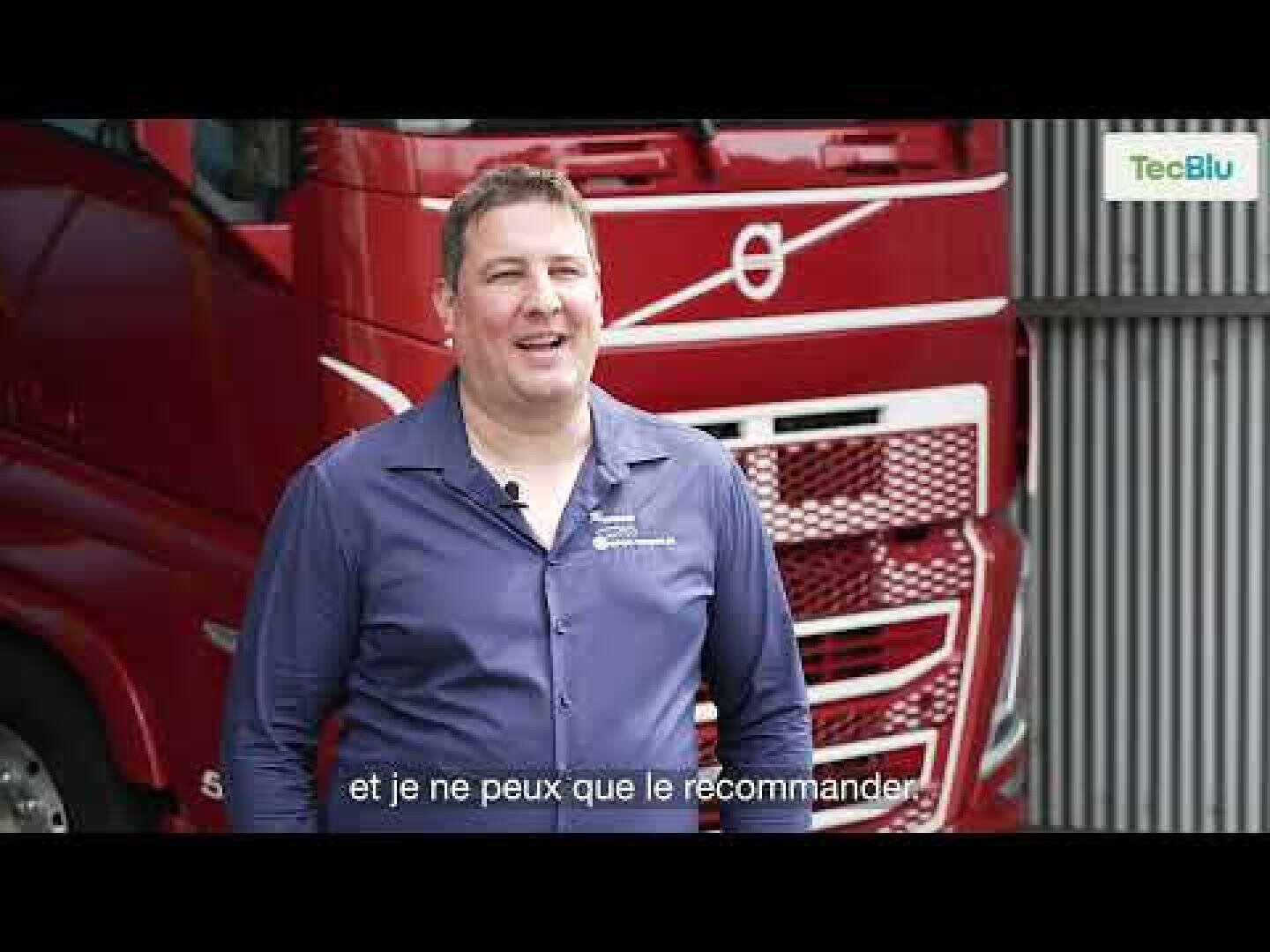 Un homme en chemise bleue se tient debout et sourit devant un camion Volvo rouge. Le logo TecBlu apparaît dans le coin supérieur droit et des sous-titres en français sont visibles en bas de l'image.