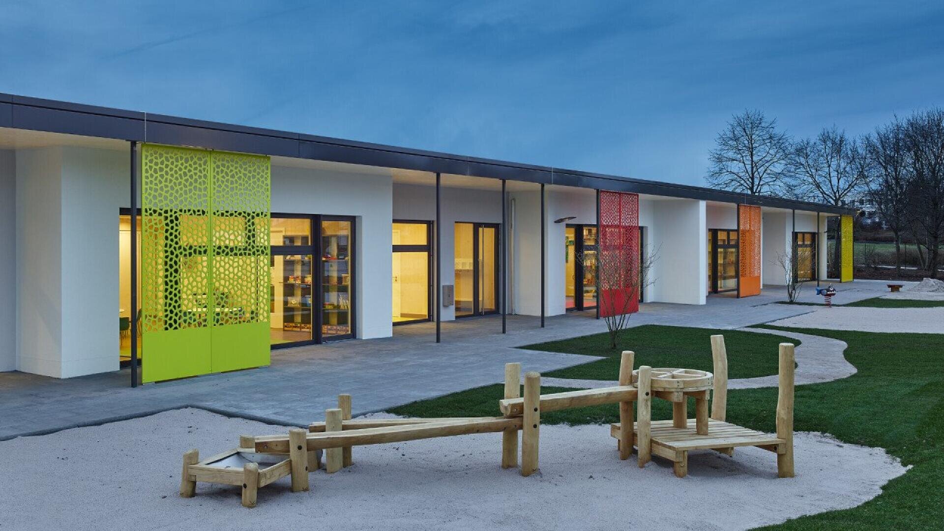 Un bâtiment moderne d'un étage avec des panneaux colorés et de grandes fenêtres se trouve derrière une aire de jeux sablonneuse avec des équipements d'escalade en bois, entourée d'herbe sous le ciel bleu du soir.