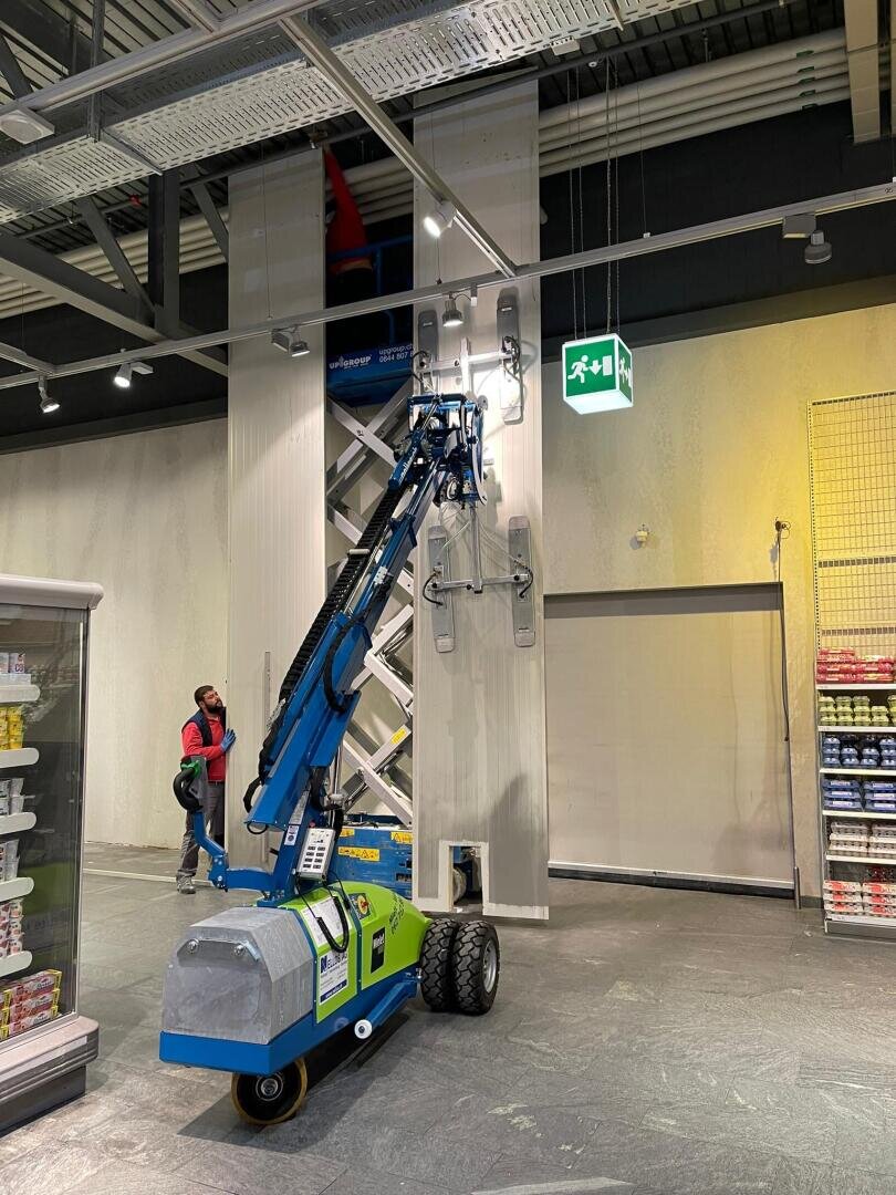 Eine blau-grüne Hebemaschine ist in einem Geschäft positioniert und hebt einen Arbeiter an, um Platten an einer hohen Wand zu installieren oder zu reparieren. Eine weitere Person steht in der Nähe von Kühlregalen. Darüber ist ein beleuchtetes Notausgangsschild zu sehen.