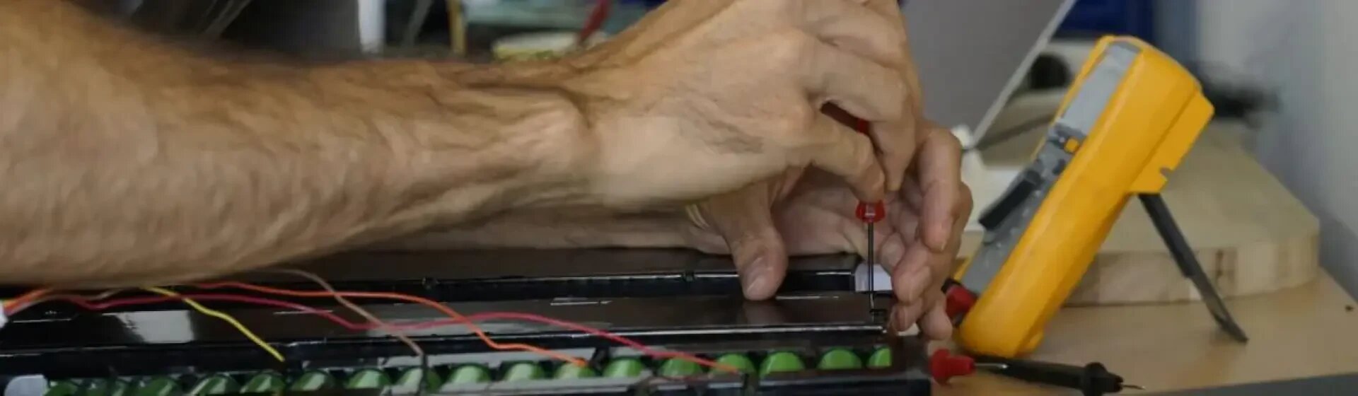 Eine Person arbeitet mit einem Schraubenzieher an einem elektronischen Gerät mit freiliegenden Batteriezellen, Drähten und einem gelben Multimeter in der Nähe auf einer Holzfläche.