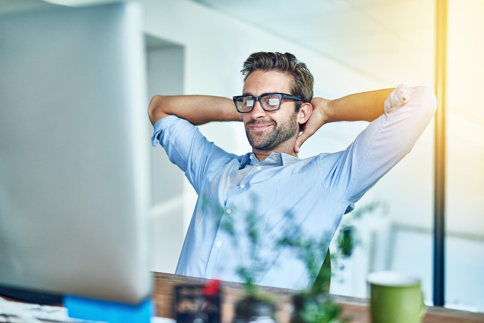 Ein Mann mit Brille und hellblauem Hemd lehnt sich in seinem Stuhl zurück, lächelt und streckt seine Hände hinter dem Kopf, während er in einem hellen Büro an einem Schreibtisch vor einem Computermonitor sitzt.
