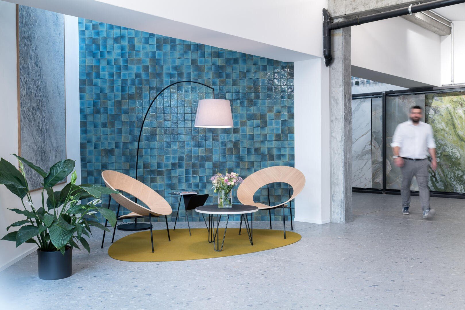 Moderne Büro-Lounge mit zwei runden Holzstühlen, einem kleinen runden Tisch mit einem Blumenstrauß, einer Stehlampe und einer Topfpflanze. Blau geflieste Akzentwand und eine Person, die im Hintergrund läuft.