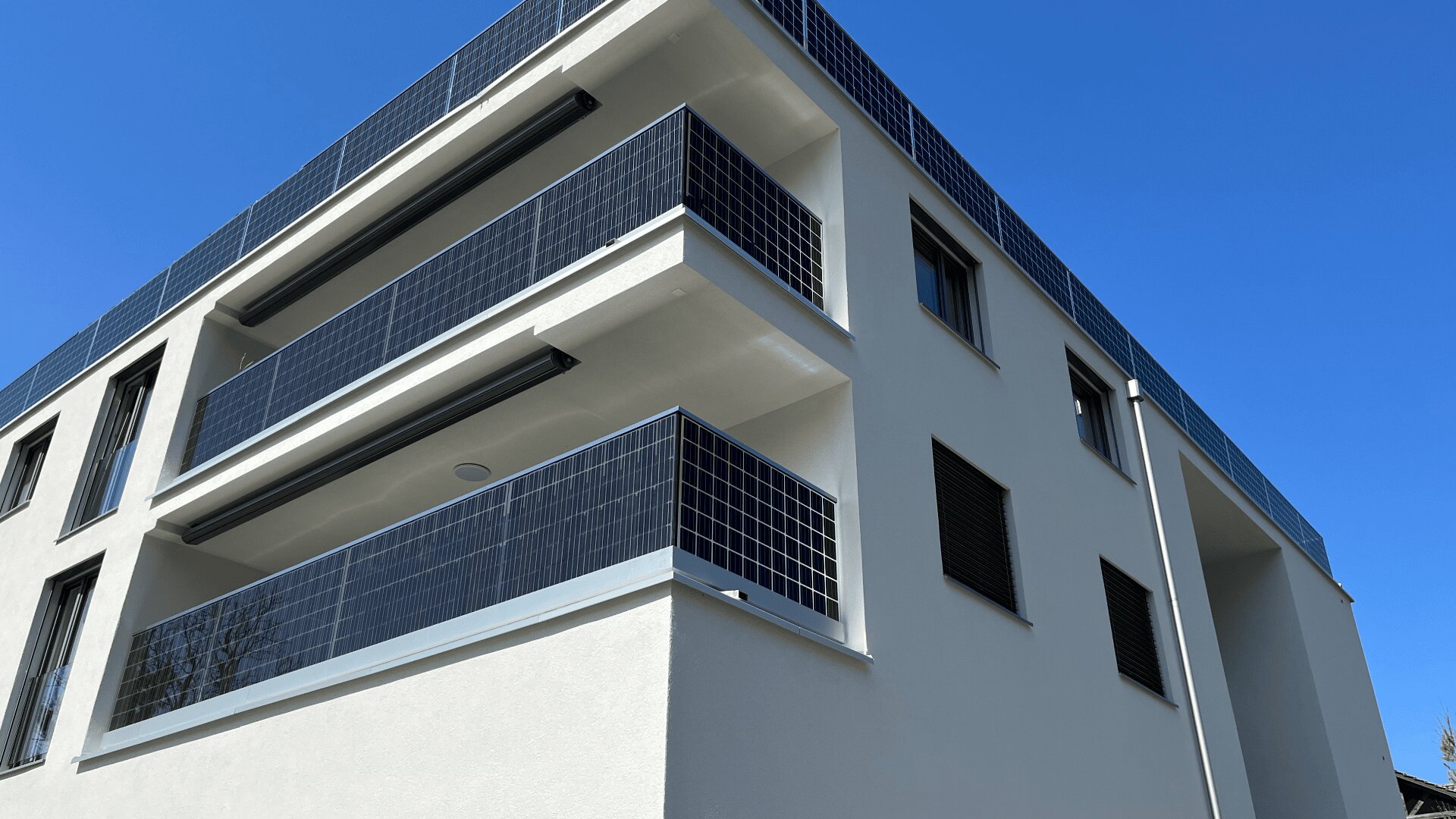 Ein modernes weißes Mehrfamilienhaus mit Balkonen, die mit schwarzen Sonnenkollektoren als Geländer und Verkleidung versehen sind, vor einem klaren blauen Himmel.