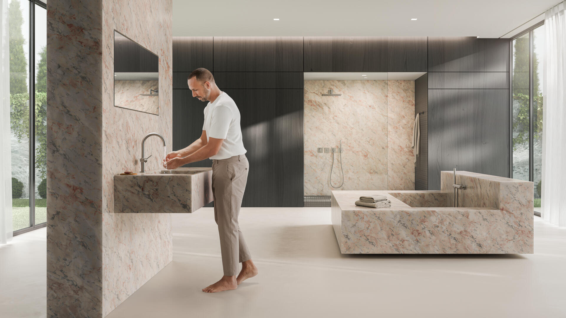 Ein Mann wäscht sich die Hände an einem Marmorwaschbecken in einem geräumigen, modernen Badezimmer mit passender Marmorbadewanne und Dusche, großen Fenstern und dunklen Holzschränken. Die Handtücher sind ordentlich auf der Wanne gestapelt.