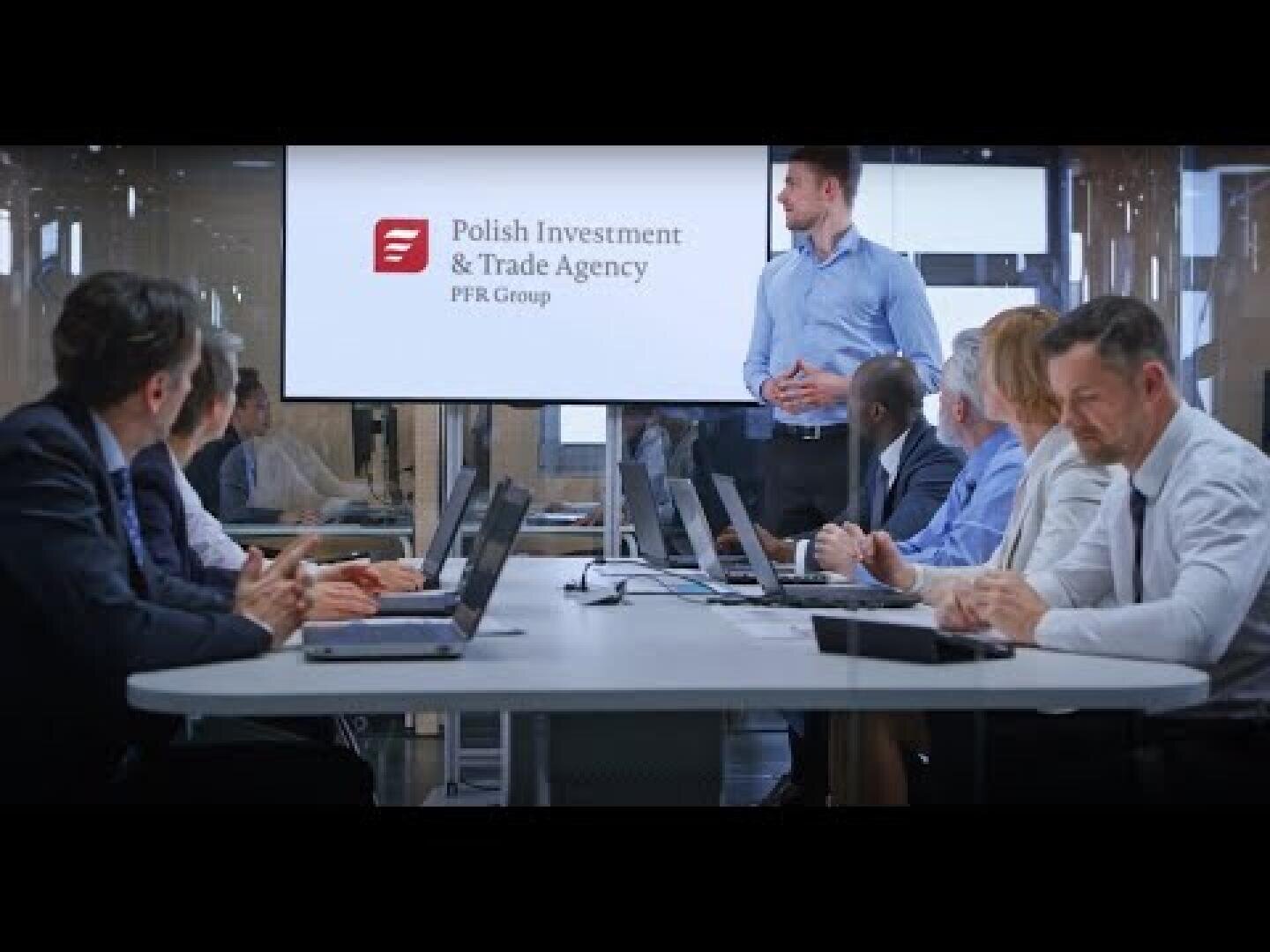 Ein Mann steht und präsentiert fünf sitzenden Kollegen in einem Konferenzraum. Sie sehen auf einen Bildschirm, auf dem die polnische Investitions- und Handelsagentur PFR Group abgebildet ist. Laptops und Papiere liegen auf dem Tisch.