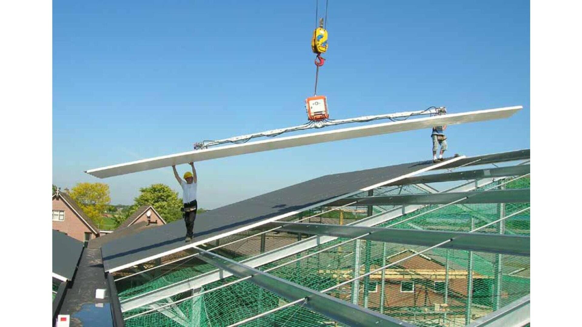 Une grue soulève un grand panneau sur le toit d'un bâtiment en construction, tandis que deux ouvriers le guident pour le mettre en place au sommet d'une charpente métallique, sous un ciel bleu dégagé.