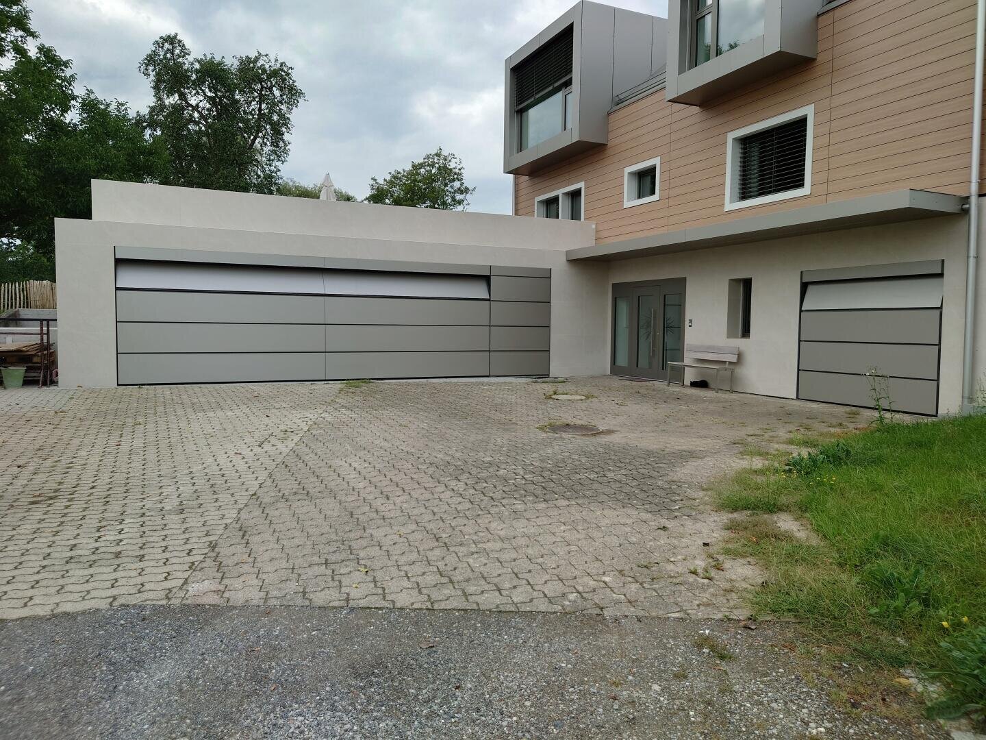 Modernes Haus mit hellgrauer Außenseite, großen Fenstern, zwei breiten Garagentoren mit horizontalen Paneelen und einer gepflasterten Einfahrt. Ein kleines Stück Gras ist auf der rechten Seite neben der Einfahrt zu sehen.