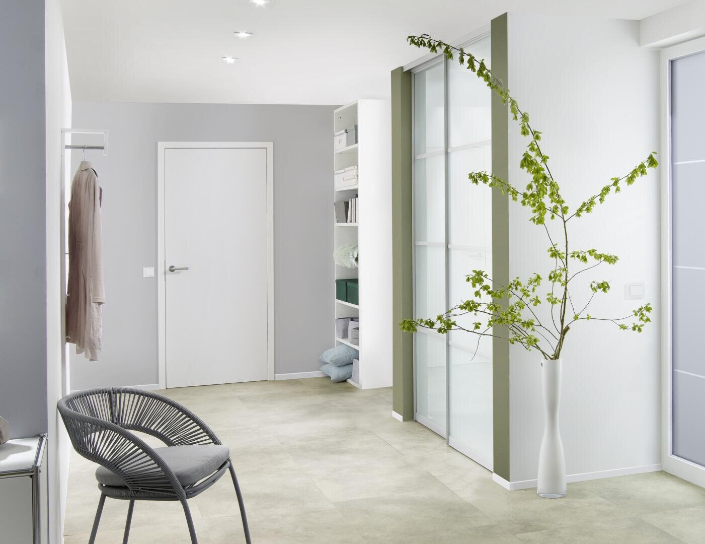 Ein moderner, minimalistischer Flur mit hellen Wänden und Böden, einem grauen Stuhl, einer hohen weißen Vase mit einem grünen Zweig, Einbauregalen mit ordentlich gefalteter Wäsche und einer geschlossenen weißen Tür.