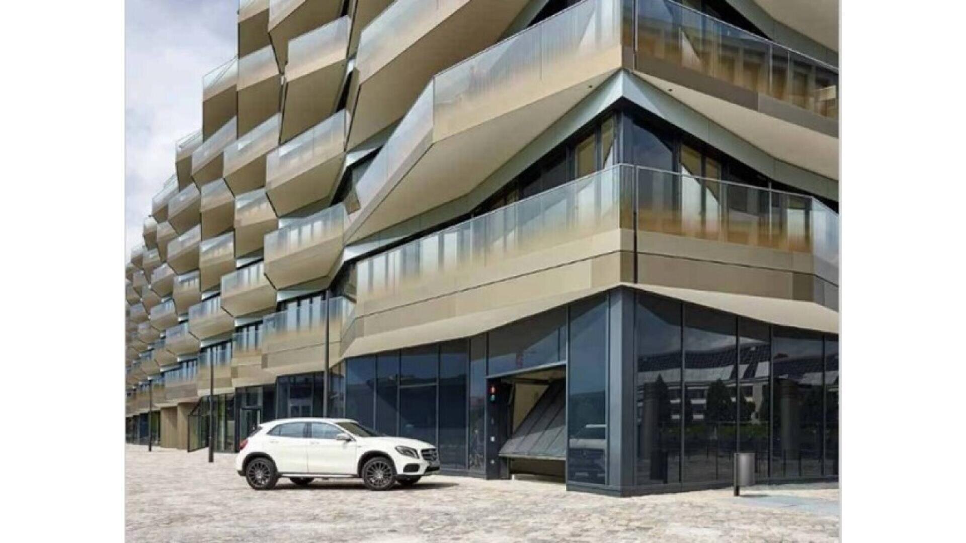 Un bâtiment moderne avec des balcons géométriques et angulaires et de grandes fenêtres en verre se trouve à côté d'un SUV blanc garé sur une zone pavée. La façade du bâtiment présente des surfaces métalliques et des lignes nettes.