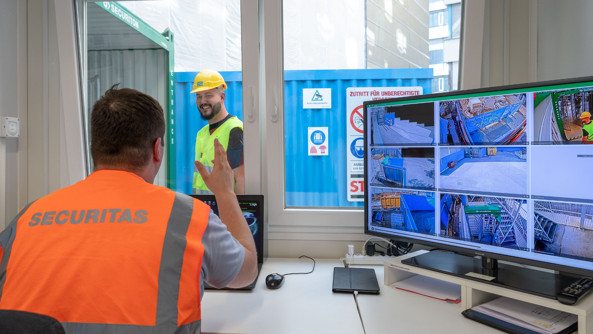 Ein Sicherheitsbeamter in einer SECURITAS-Weste überwacht die CCTV-Bildschirme in einem Büro und gestikuliert in Richtung eines Bauarbeiters in einer gelben Weste und mit Helm, der draußen an einem Container steht.