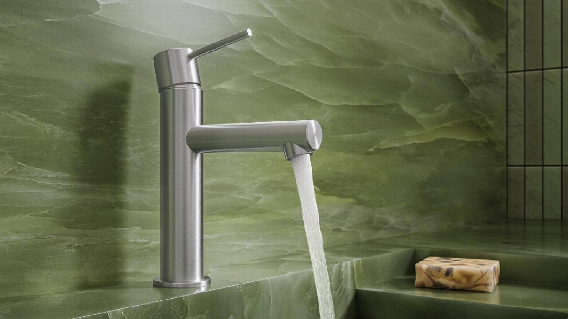 Un robinet moderne en acier inoxydable avec de l'eau qui s'écoule est monté sur un comptoir en marbre vert, à côté d'une barre de savon. L'arrière-plan est composé de marbre vert assorti et de lignes verticales de carrelage noir.