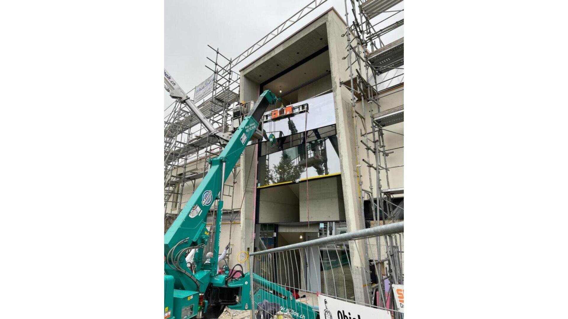 Une grue verte soulève un grand panneau de verre à installer au deuxième étage d'un bâtiment moderne en construction, entouré d'échafaudages et de barrières de sécurité.