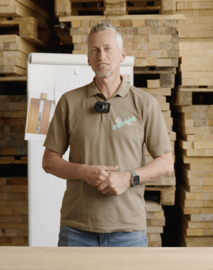 Un homme vêtu d'un polo marron se tient devant des planches de bois empilées. Il a les cheveux gris courts, une barbe et porte une smartwatch et un microphone. En arrière-plan, on peut voir un tableau blanc.