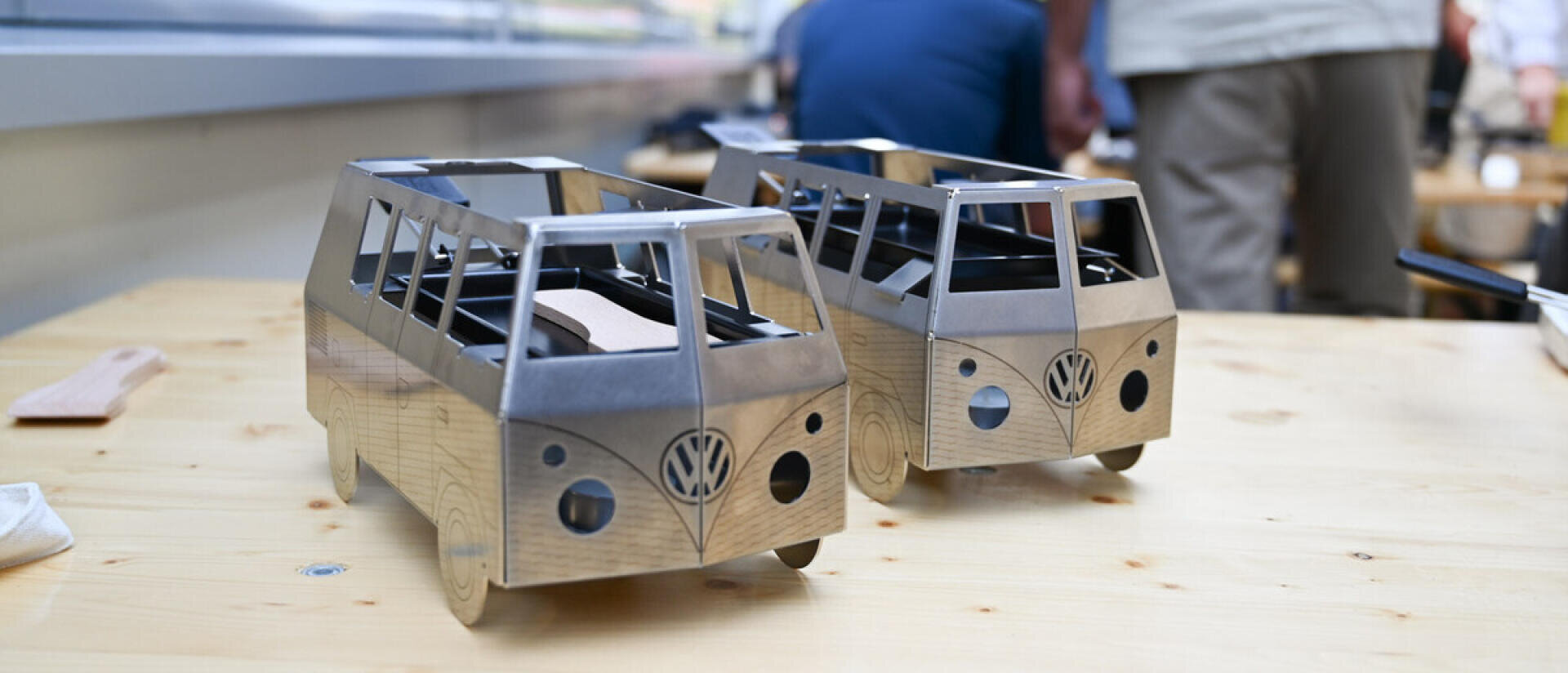 Auf einem hellen Holztisch stehen zwei kleine Metallmodelle, die an einen VW-Bus erinnern; im Hintergrund stehen und arbeiten Menschen, die unscharf sind.