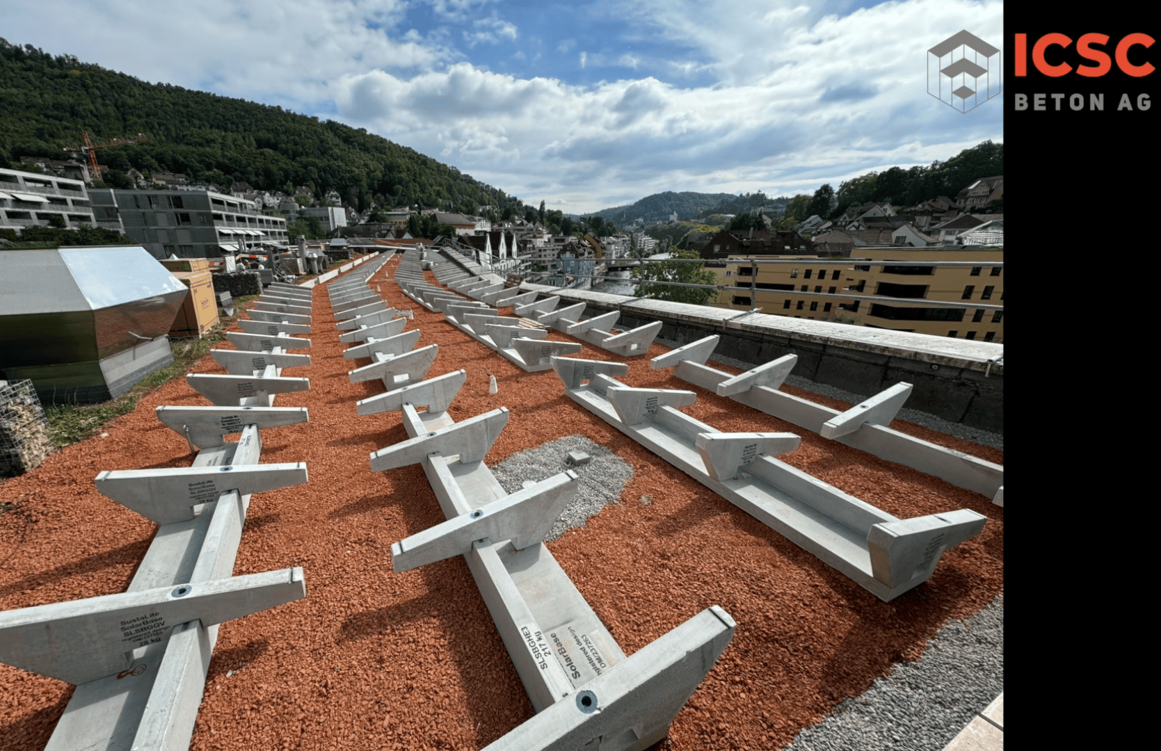 Auf einem mit rötlichem Kies bedeckten Dach sind Reihen von großen Betonstützen angeordnet, die eine hügelige Stadtlandschaft unter einem teilweise bewölkten Himmel überblicken. In der rechten oberen Ecke erscheint das Logo der ICSC BETON AG.