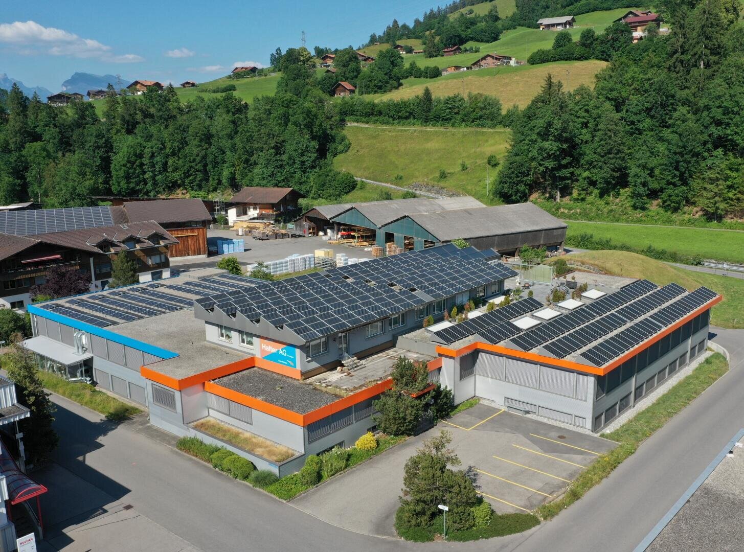 Ein modernes Industriegebäude mit Sonnenkollektoren auf dem Dach, umgeben von Grün, Hügeln und Bäumen, mit Häusern in der Nähe und einem klaren blauen Himmel im Hintergrund.