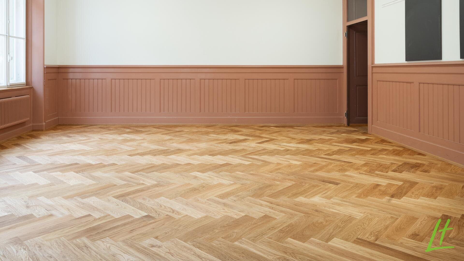 Ein leerer Raum mit hellem Fischgrätparkett, pfirsichfarbener Vertäfelung und Verkleidung, weißen oberen Wänden, einem großen Fenster auf der linken Seite und einer geschlossenen Tür an der gegenüberliegenden Wand.