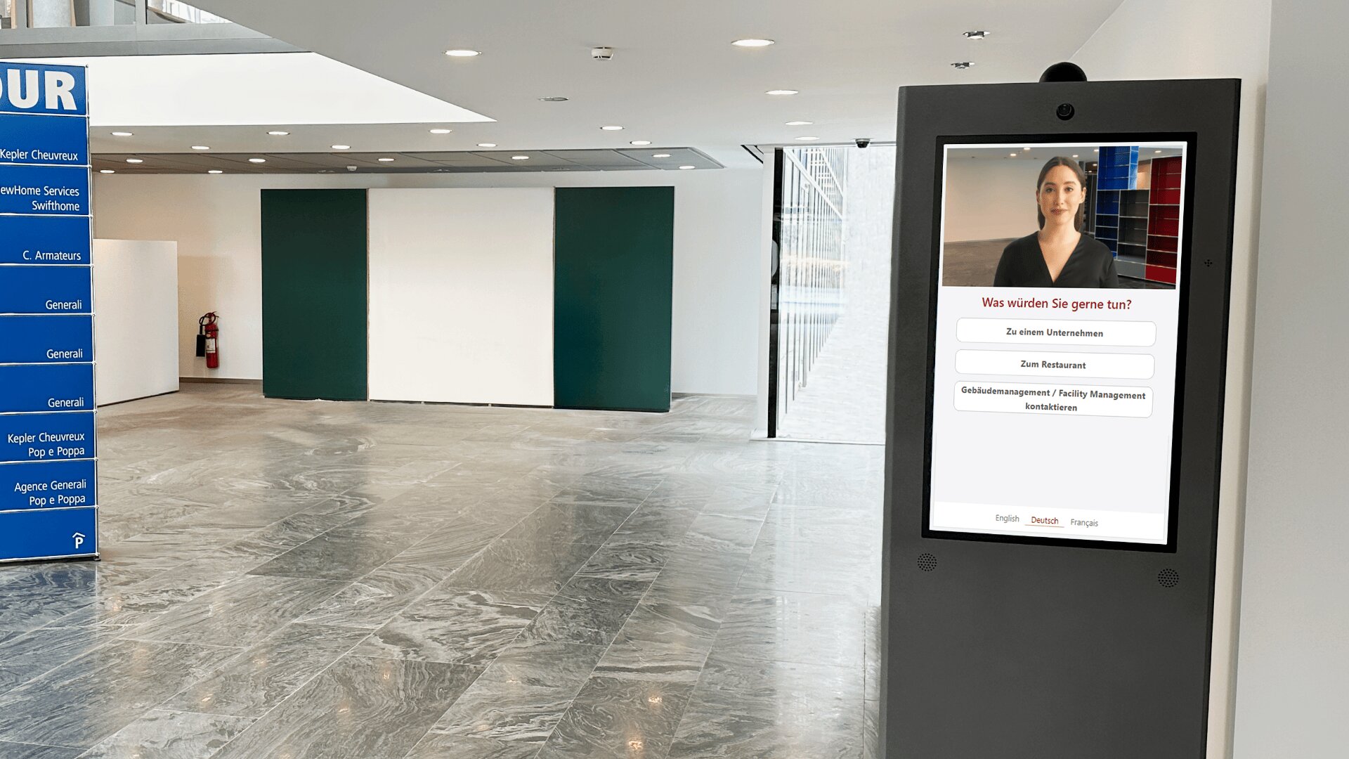 Ein digitaler Informationskiosk mit einer Touchscreen-Oberfläche steht in einem modernen Gebäude mit Marmorfußboden. Der Bildschirm des Kiosks zeigt das Foto einer Frau und deutsche Menüoptionen. Auf der linken Seite ist ein blaues Telefonbuchschild zu sehen.
