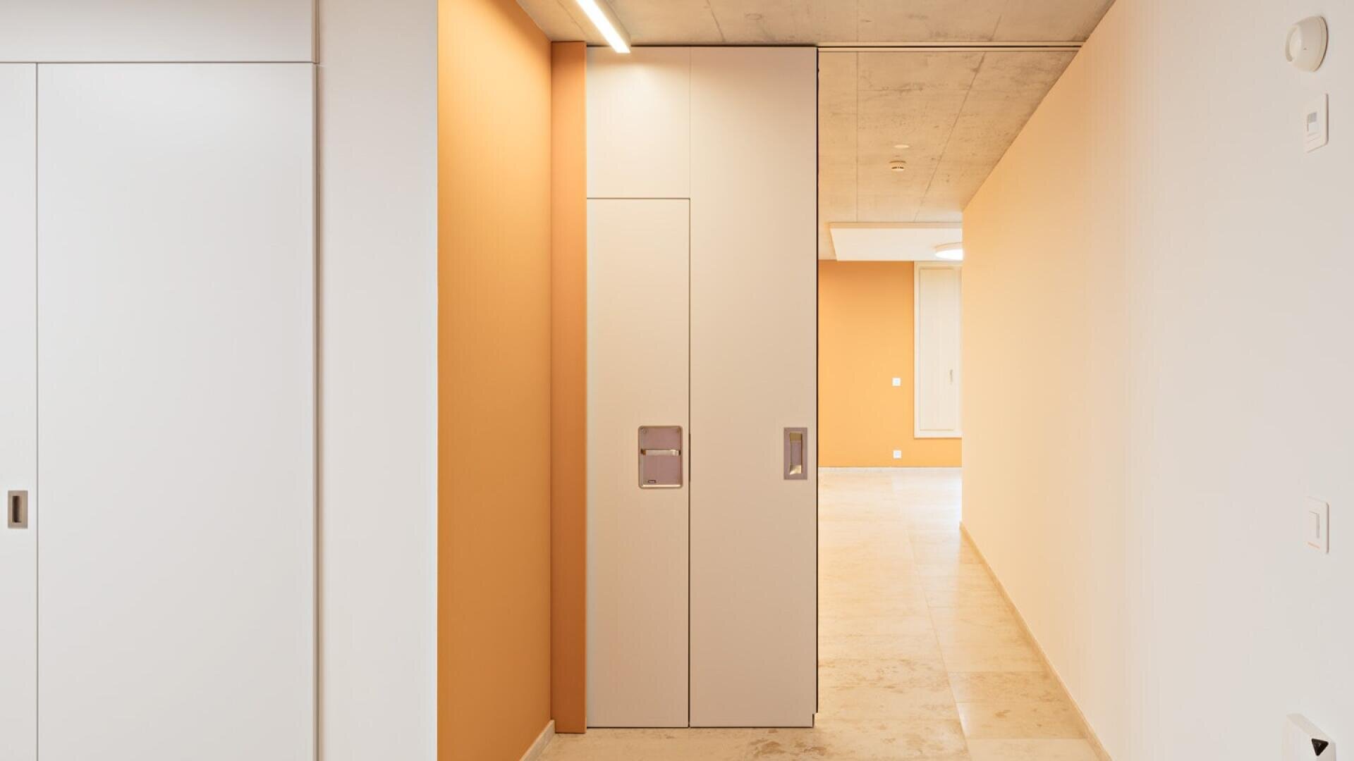 Un couloir moderne et minimaliste avec des carreaux de sol beiges, des murs orange et blancs, un éclairage encastré et d'élégantes portes coulissantes blanches. Le design est propre et clair et crée une atmosphère lumineuse et ouverte.