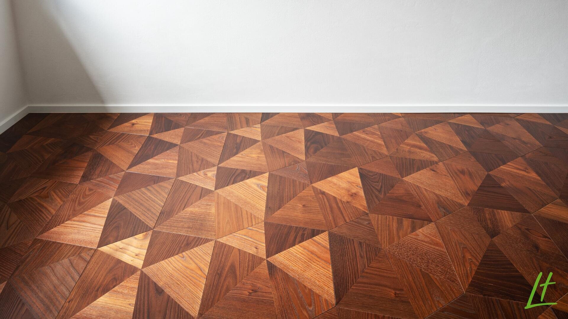 Parquet géométrique en bois avec un motif de triangles et de losanges dans différentes teintes de bois, à côté d'un mur blanc uni et d'un coin de la pièce.