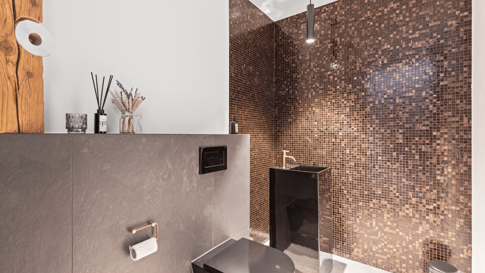 Modernes Badezimmer mit dunkel gefliesten Wänden und Mosaik-Akzenten in der Dusche, mit einer schwarzen Toilette, einem Regal mit Dekorationsartikeln und einem goldenen Wasserhahn. Eine Rolle Toilettenpapier ist an einem rustikalen Holzbalken befestigt.