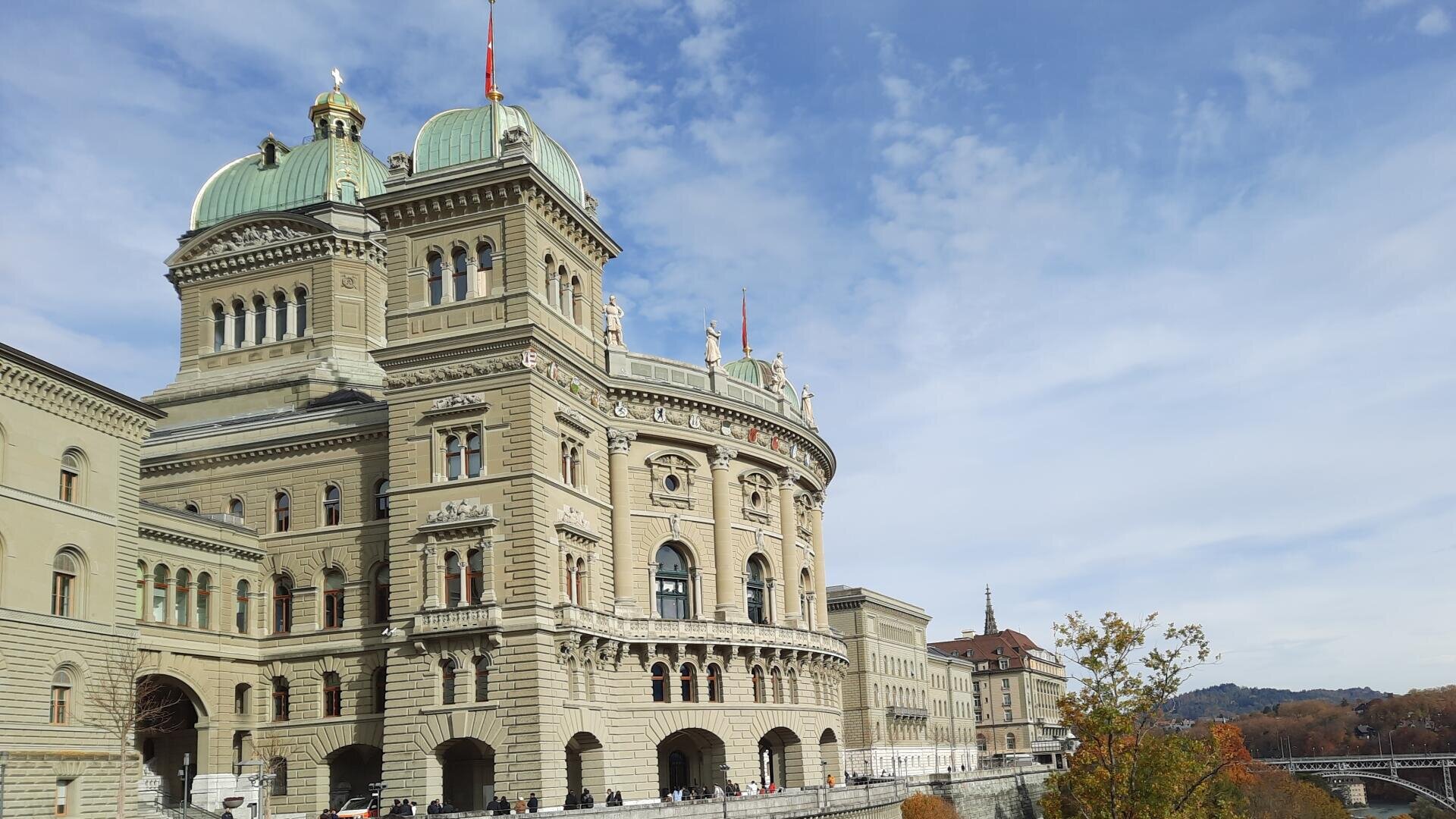 Das Bild zeigt das Bundeshaus in Bern, Schweiz, mit seinen charakteristischen grünen Kuppeln und seiner kunstvollen Architektur unter einem teilweise bewölkten Himmel. Menschen gehen die Straße entlang, und im Hintergrund sind Herbstbäume zu sehen.