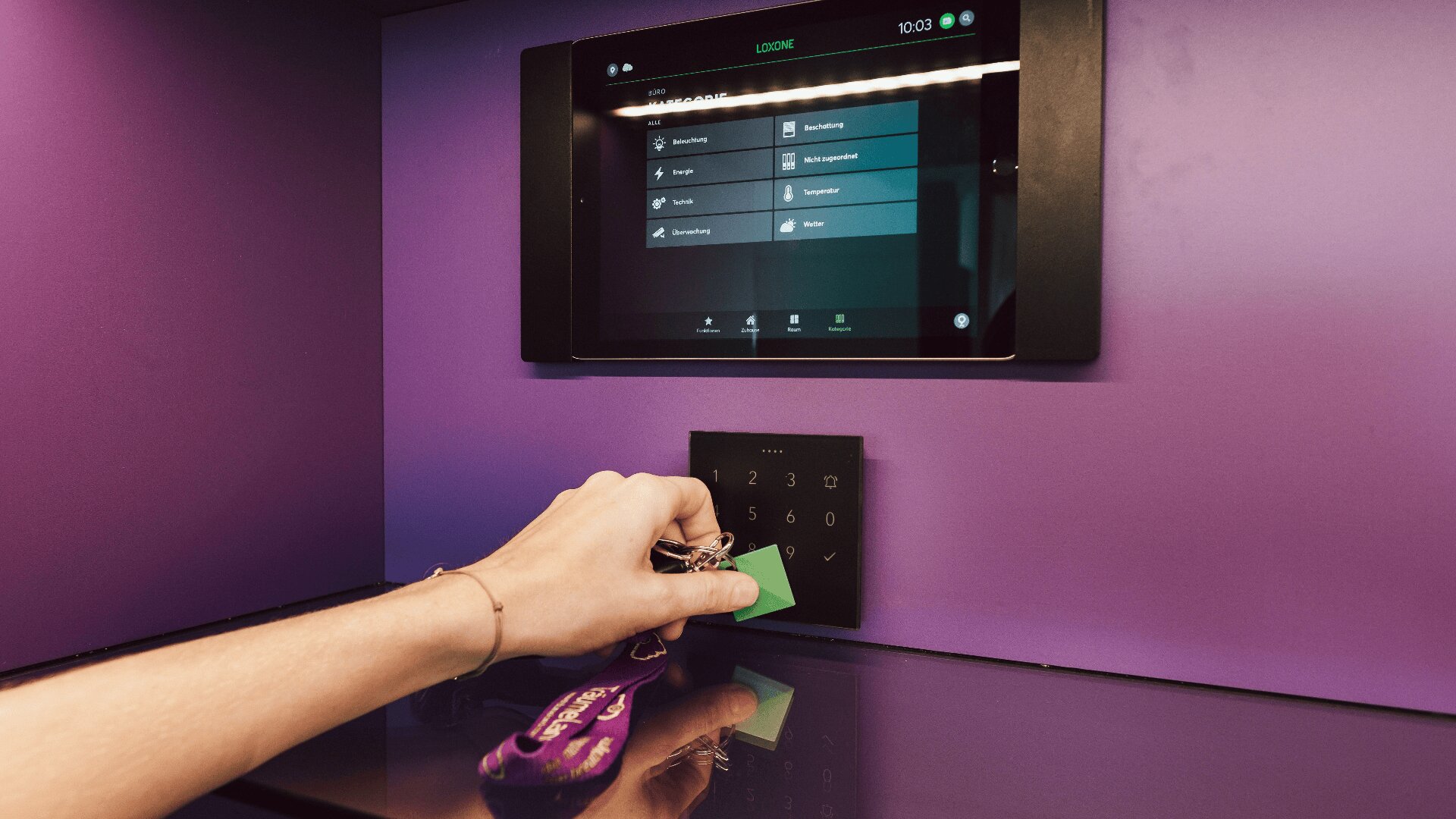Eine Person hält eine grüne Schlüsselkarte und einen Schlüsselbund in der Hand und scannt sie auf einem schwarzen Tastenfeld unter einem an einer lila Wand angebrachten Touchscreen-Bedienfeld.