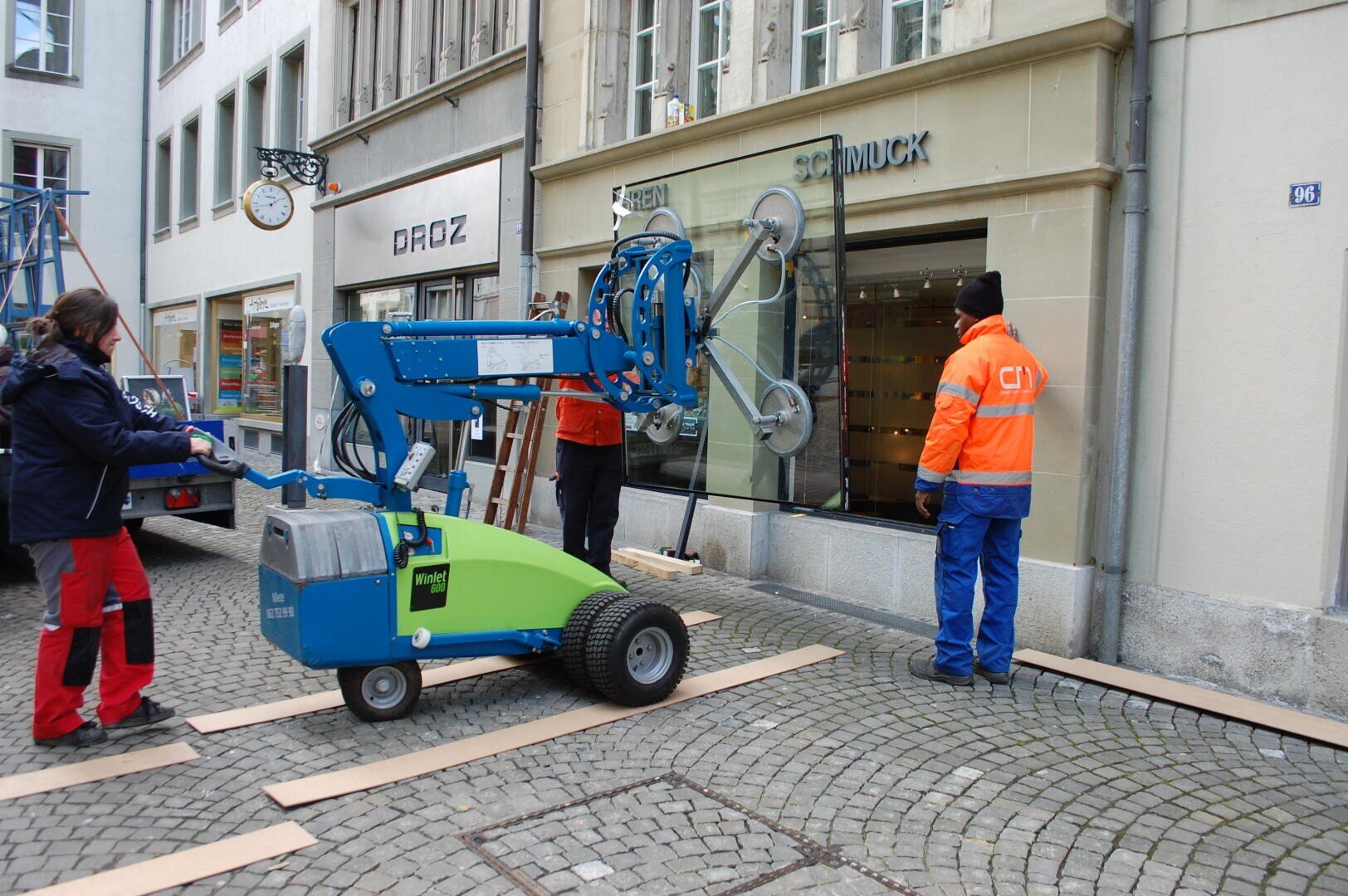 Drei Arbeiter setzen mit einer blaugrünen Glasbearbeitungsmaschine eine große Fensterscheibe in einem Geschäft mit der Aufschrift Schmuck in einer Kopfsteinpflasterstraße ein. Holzbretter bedecken den Gehweg zum Schutz.