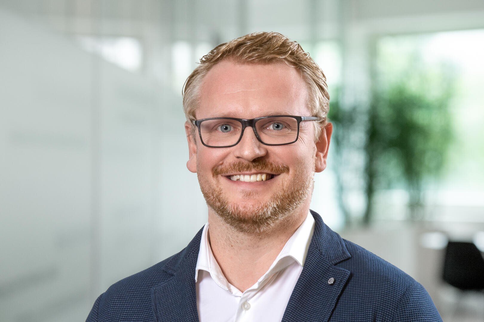 Un homme souriant aux cheveux blonds courts, portant une barbe et des lunettes, vêtu d'une chemise blanche et d'un blazer bleu, se tient dans un bureau moderne et lumineux, avec des plantes vertes floues en arrière-plan.