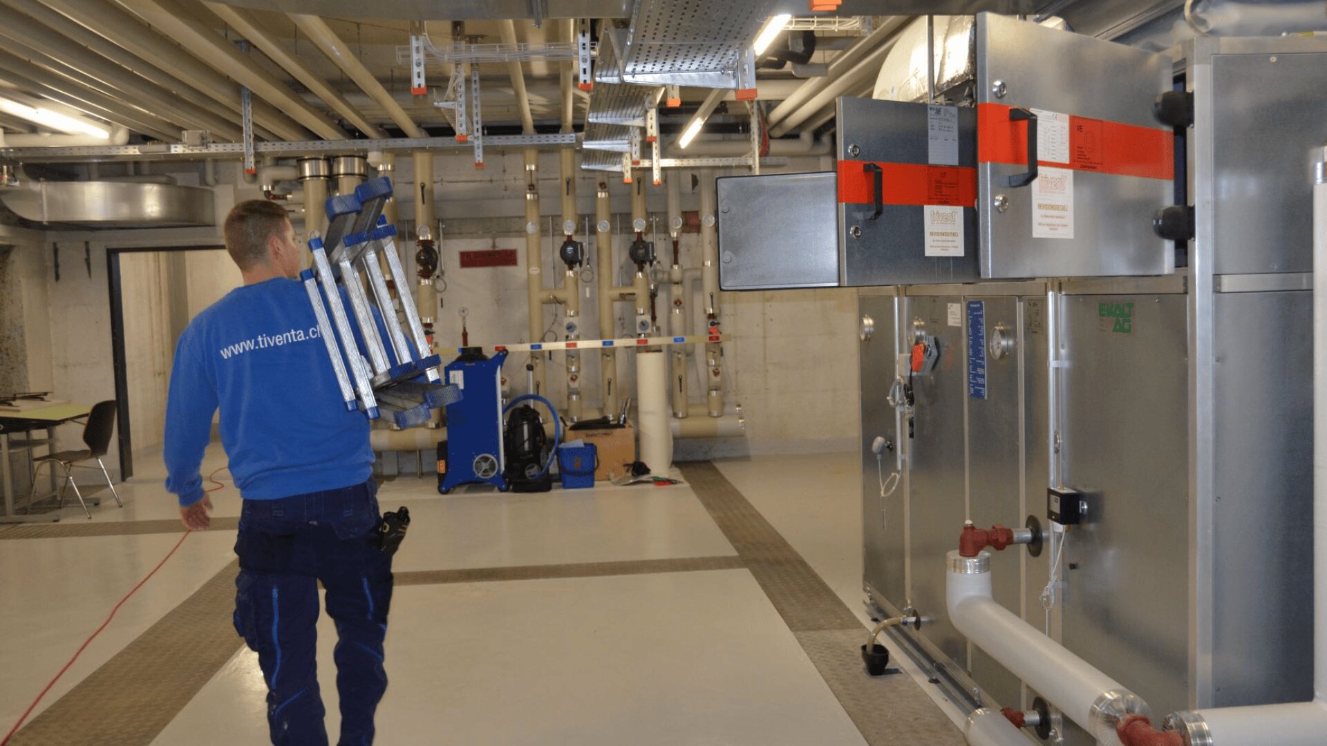 Ein Arbeiter in einem blauen Hemd trägt eine zusammengeklappte Leiter in einer Industrieanlage mit freiliegenden Rohren, großen Metallgeräten und einem Tisch mit Stühlen im Hintergrund.