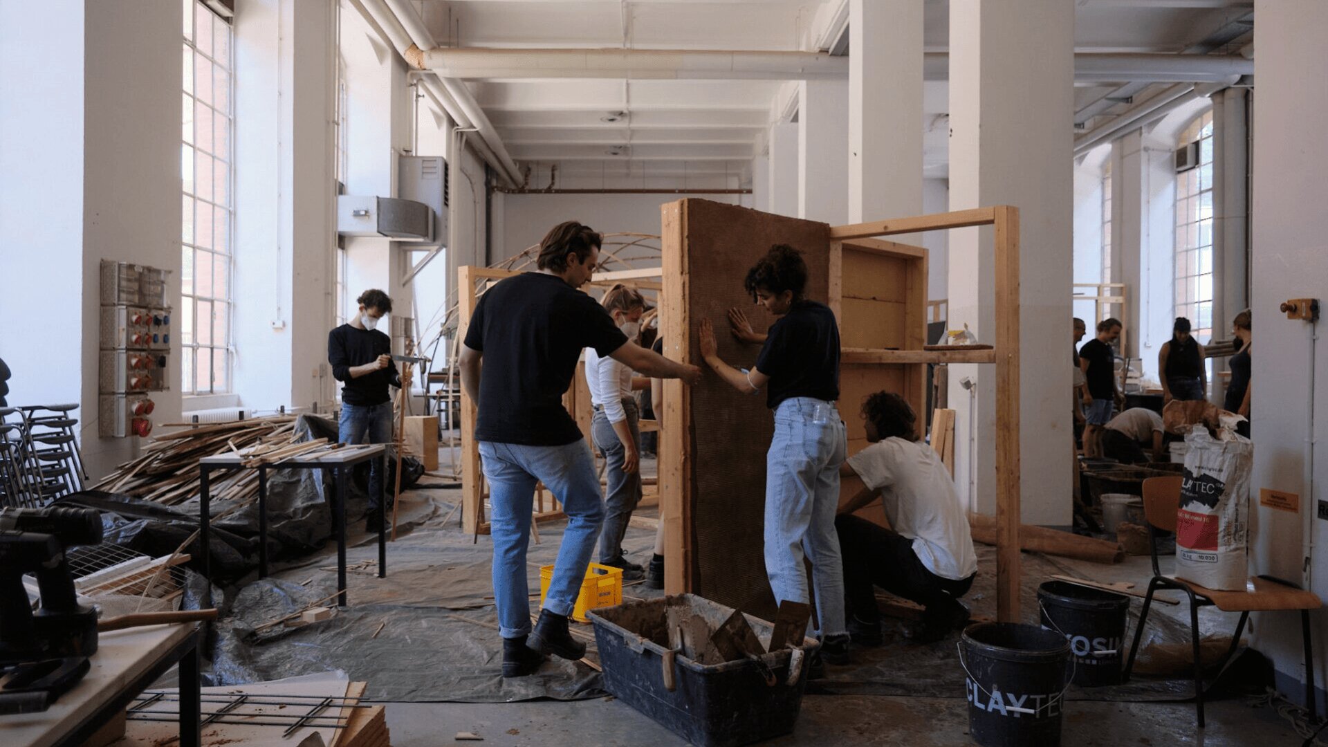 In einer geräumigen, gut beleuchteten Werkstatt arbeiten mehrere Personen zusammen, setzen Holzstrukturen zusammen und hantieren mit Baumaterialien. Einige schleifen oder reinigen Holzplatten, während andere in der Nähe knien oder stehen und zusammenarbeiten.