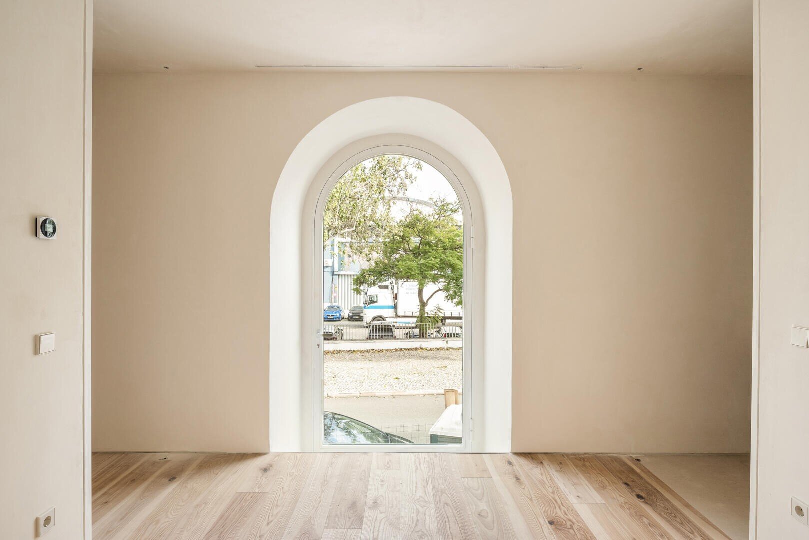 Ein minimalistischer Raum mit hellen Holzböden und beigen Wänden verfügt über ein großes Bogenfenster, das einen Blick auf die Bäume, die geparkten Autos und die Gebäude im Freien bietet.
