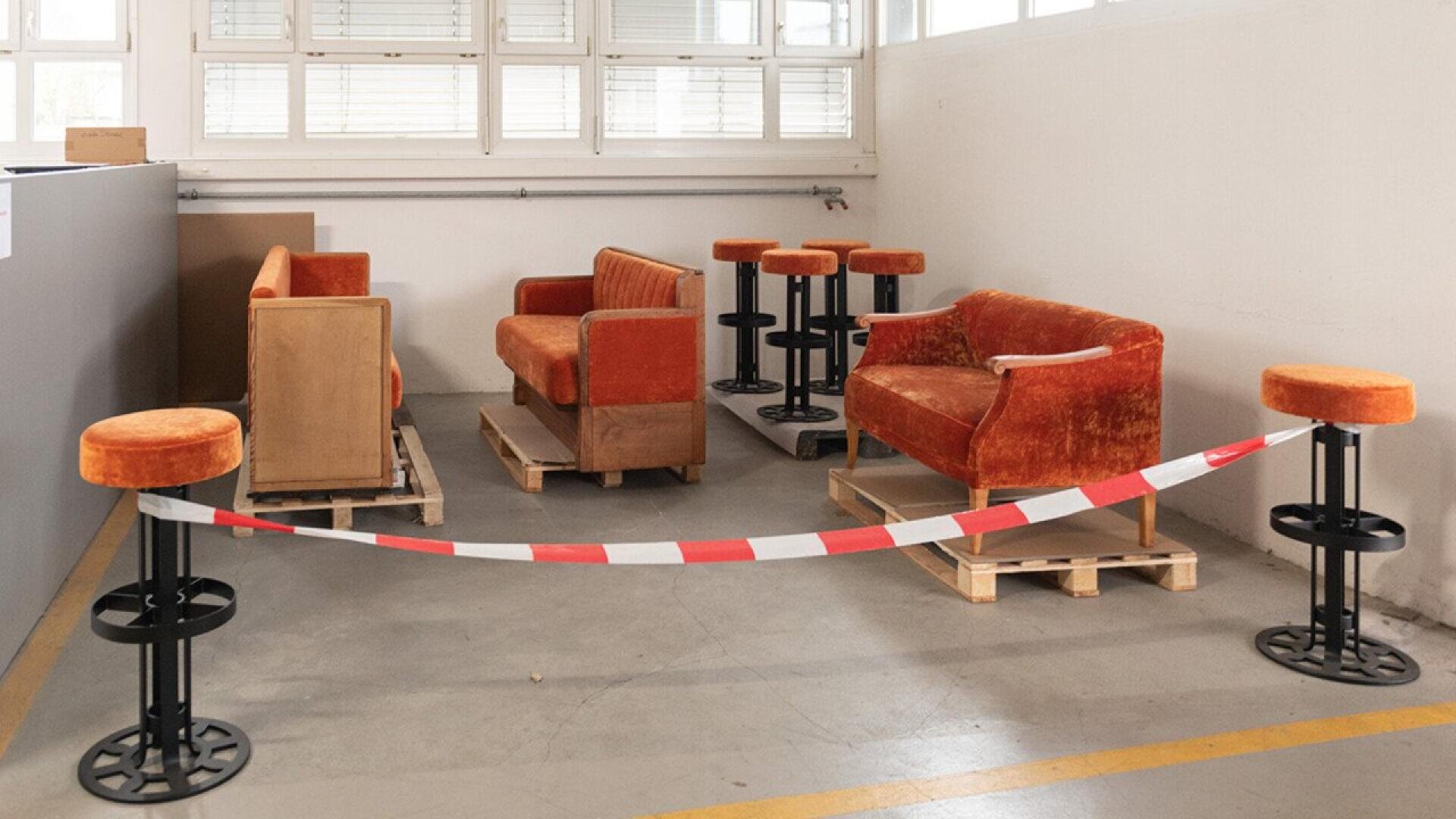 Plusieurs canapés et tabourets en velours orange sont posés sur des palettes en bois dans un coin d'une pièce, dont l'accès est bloqué par un ruban adhésif rayé rouge et blanc. La pièce est dotée de grandes fenêtres et d'un sol en béton.