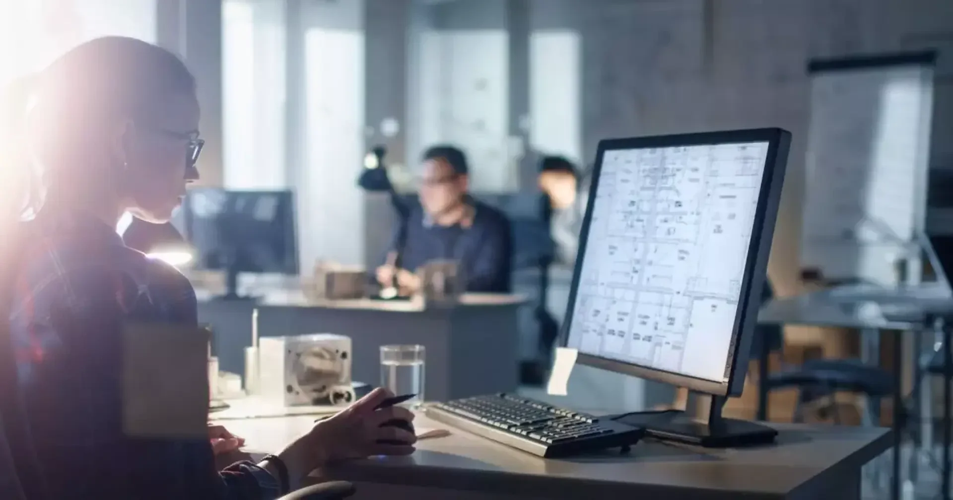 Eine Person, die an einem Schreibtisch in einem schwach beleuchteten Büro arbeitet und architektonische Entwürfe oder technische Zeichnungen auf einem großen Computermonitor betrachtet, wobei die Mitarbeiter im Hintergrund zu sehen sind.
