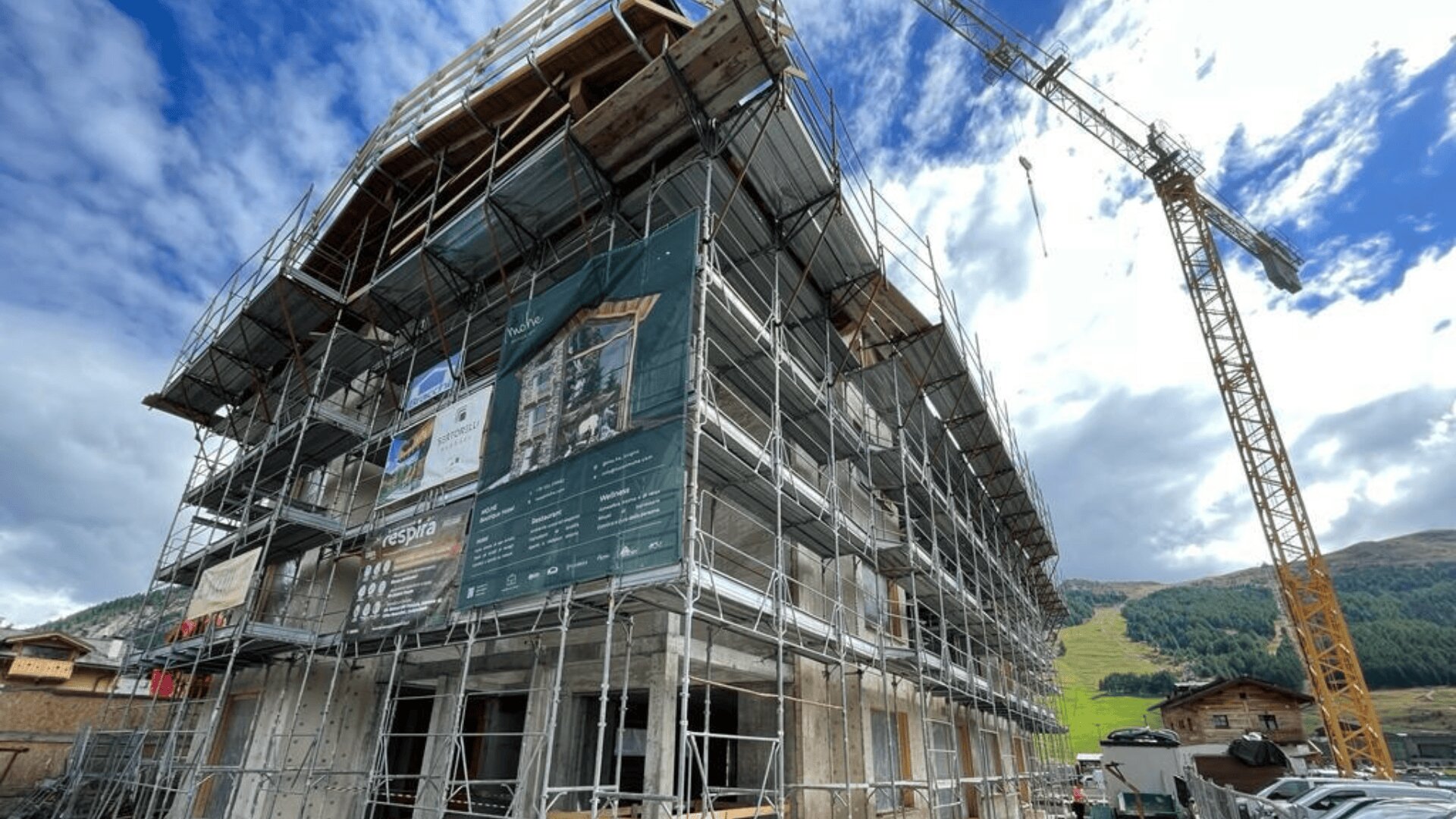 Un bâtiment de plusieurs étages en construction est entouré d'échafaudages, avec des banderoles sur un côté. Une grande grue jaune se trouve à proximité, et des collines verdoyantes avec quelques maisons sont visibles à l'arrière-plan sous un ciel partiellement nuageux.