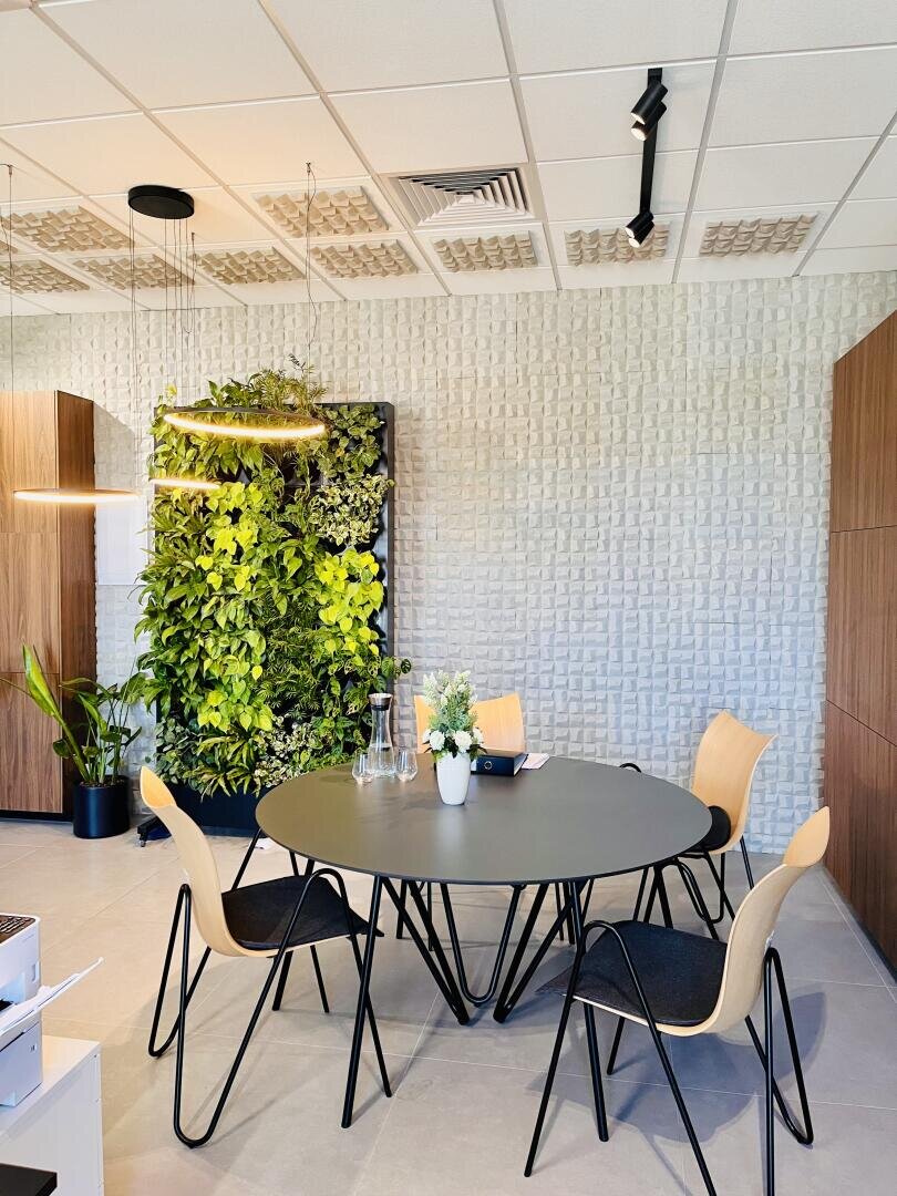 Une salle de réunion moderne avec une table ronde noire, quatre chaises en bois, des plantes sur la table, des armoires en bois, un jardin vertical vert sur le mur et des panneaux muraux géométriques sous un éclairage lumineux.