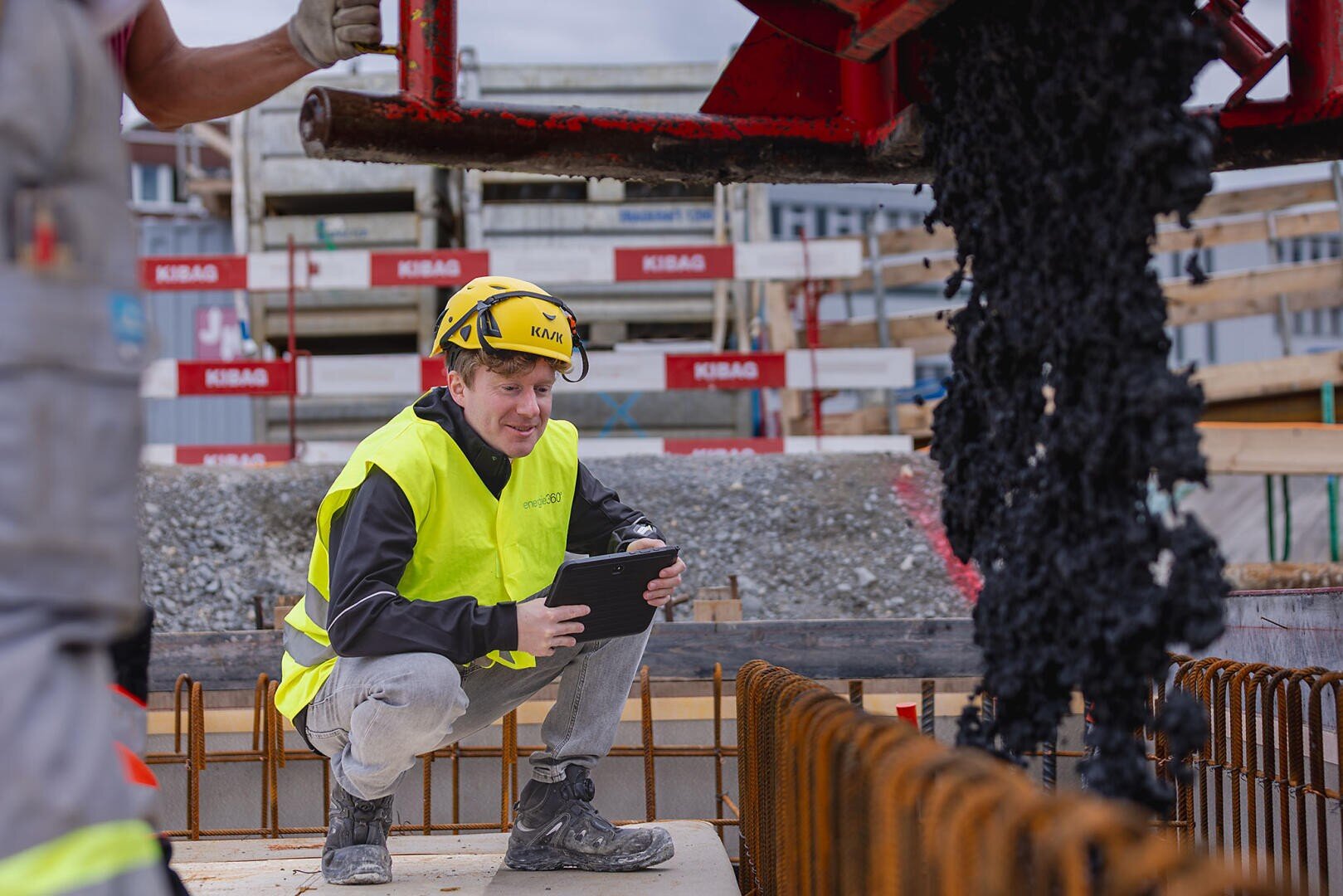 Un ouvrier du bâtiment, portant un casque jaune et un gilet de sécurité, est accroupi avec une tablette et observe comment l'asphalte est coulé sur un chantier, tandis qu'en arrière-plan, on voit des fers à béton et des matériaux de construction.