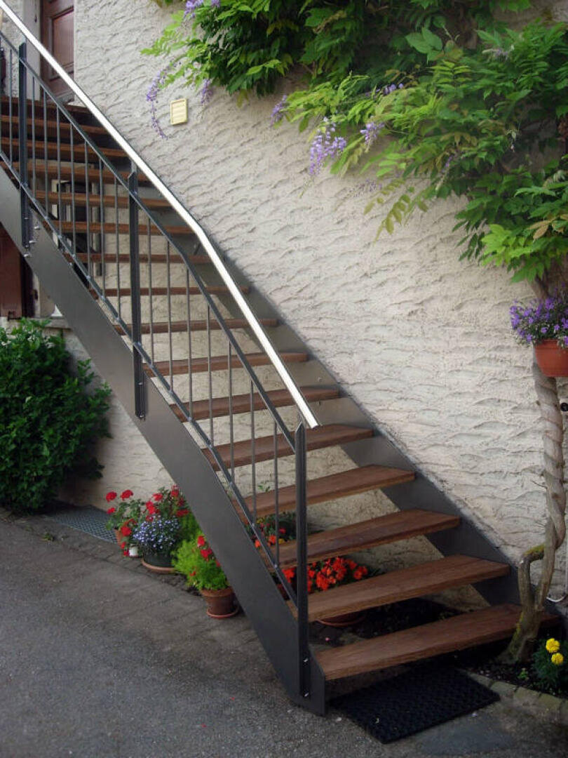 Eine Außentreppe aus Metall und Holz, die an einer beigen Stuckwand befestigt ist, umgeben von Topf- und Kletterpflanzen, darunter rote, violette und weiße Blumen. Die Treppe führt zu einem oberen Durchgang.