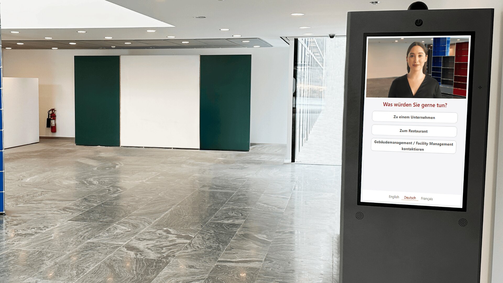 Un kiosque à écran tactile se trouve dans un hall d'entrée moderne et vide, au sol de marbre gris. L'écran affiche l'image d'une femme et des options en allemand, proposant la gestion des visiteurs et des indications sur l'établissement.