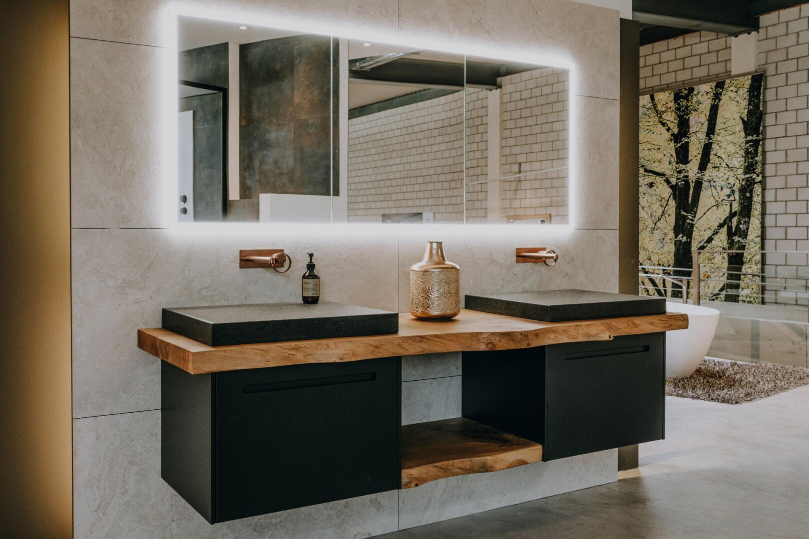 Modernes Badezimmer mit einem großen, hinterleuchteten Spiegel, zwei schwarzen Waschbecken auf einer hölzernen Arbeitsplatte, Wandarmaturen und dekorativen Gegenständen, mit minimalistischem Design und neutralen Farben.