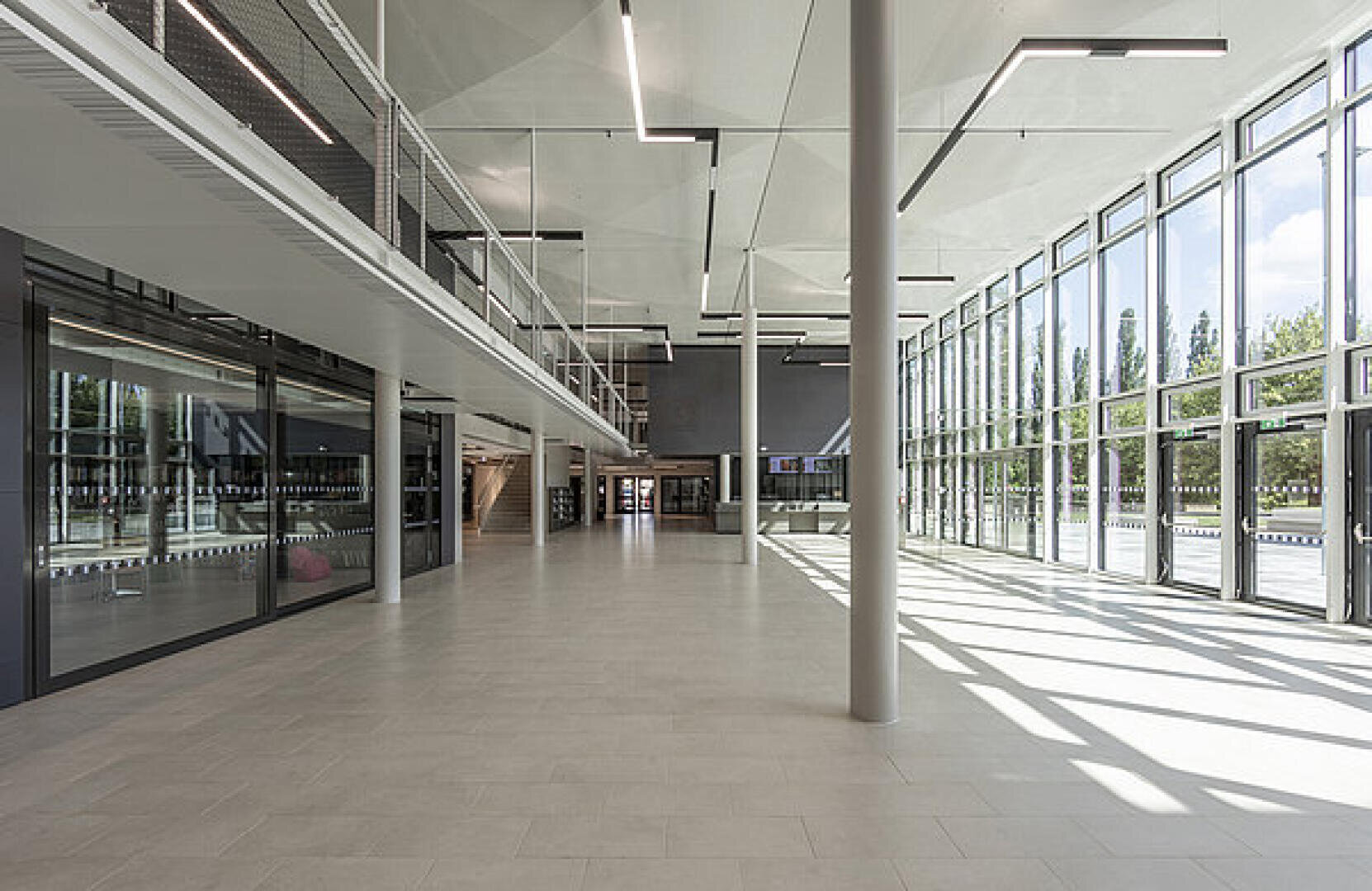 Ein geräumiges, modernes Gebäudeinneres mit großen, raumhohen Fenstern, natürlichem Licht, gefliesten Böden und einem Zwischengeschoss mit Glasgeländer. Der Raum wirkt offen und minimalistisch.