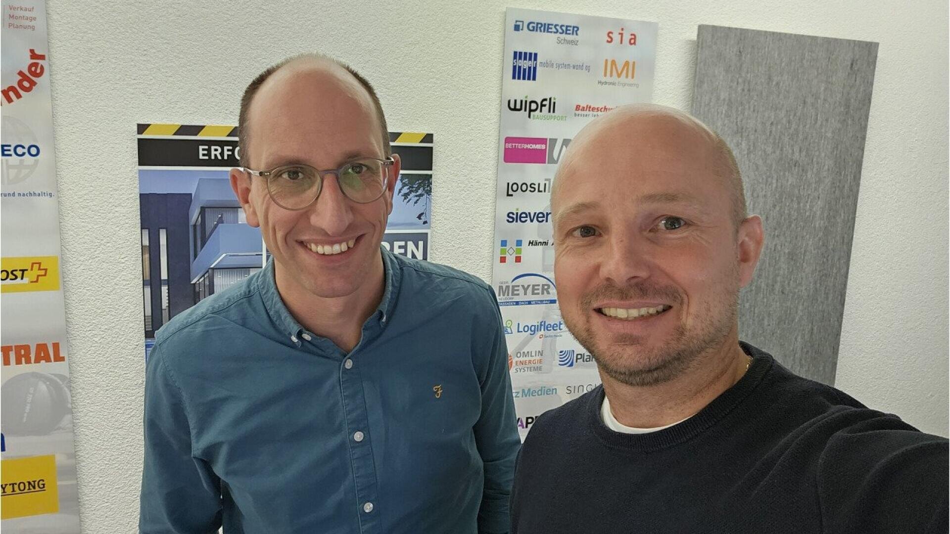 Deux hommes souriants se tiennent à l'intérieur devant un mur blanc derrière lequel on peut voir divers logos et affiches d'entreprises. L'un porte des lunettes et une chemise bleue, l'autre un pull noir.
