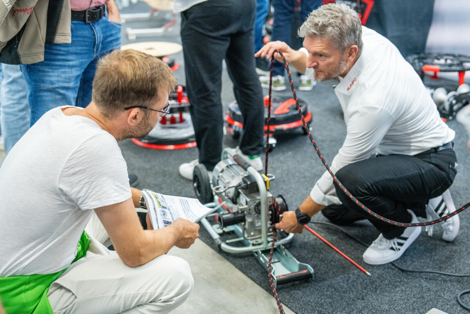 Zwei Männer begutachten bei einer Veranstaltung in einer Halle ein mechanisches Gerät, das auf dem Boden steht. Ein Mann hält Unterlagen in der Hand, während der andere kniet und die Funktionsweise des Geräts vorführt. Im Hintergrund sind weitere Maschinen und Personen zu sehen.