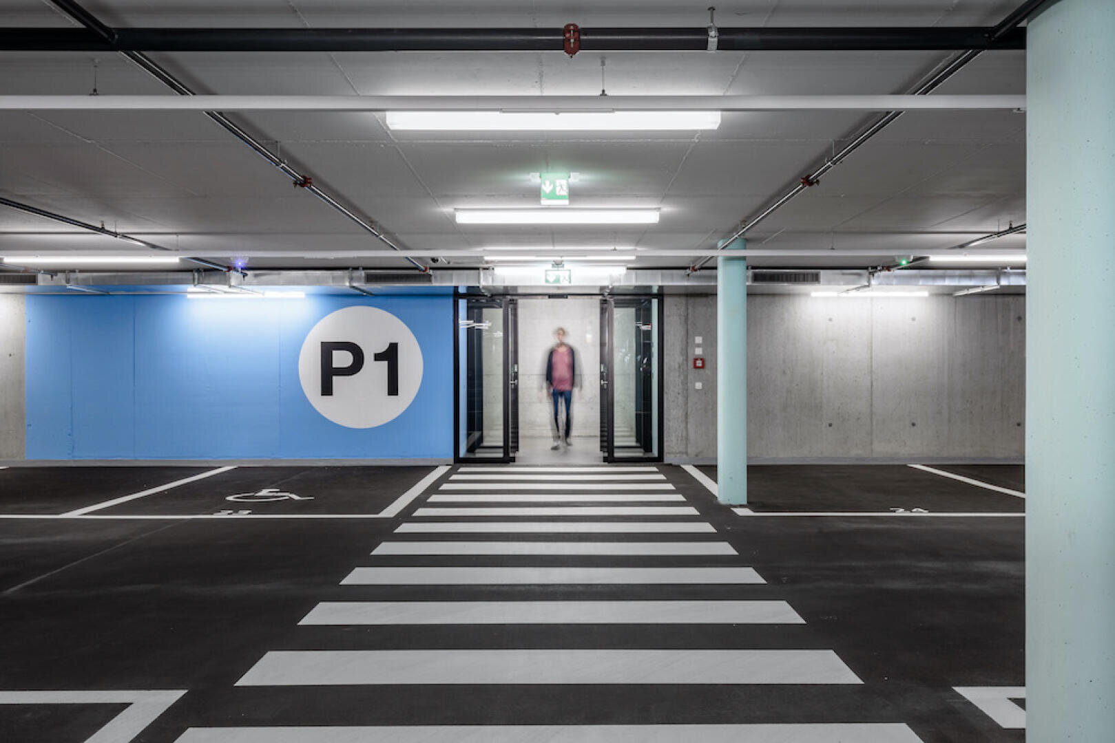Un parking souterrain moderne avec un mur bleu portant l'inscription "P1", des lignes de parking blanches, un passage pour piétons et une personne qui se dirige vers la caméra à travers des portes vitrées en arrière-plan.