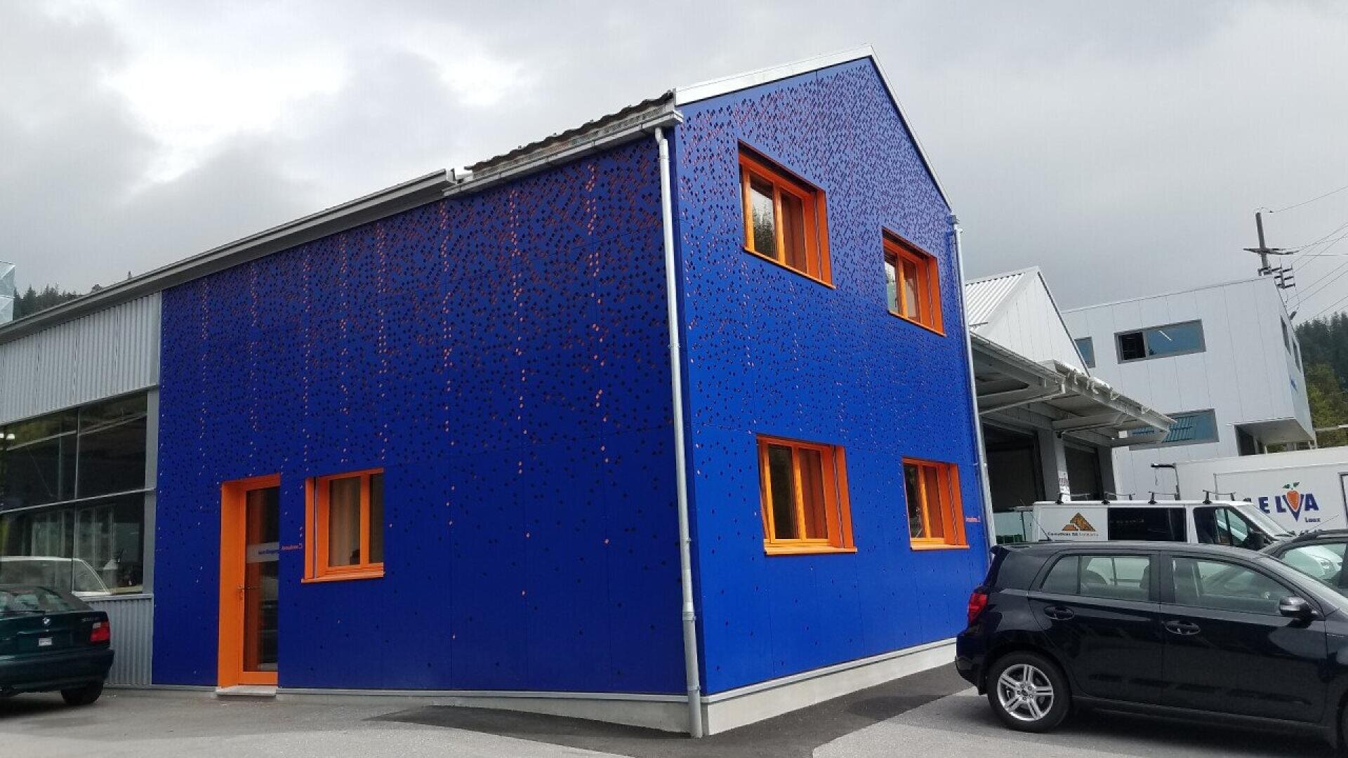Un petit bâtiment avec un mur extérieur bleu vif et des cadres de fenêtres et une porte orange vif. Le bâtiment est situé à côté de structures industrielles et de voitures garées sous un ciel nuageux.