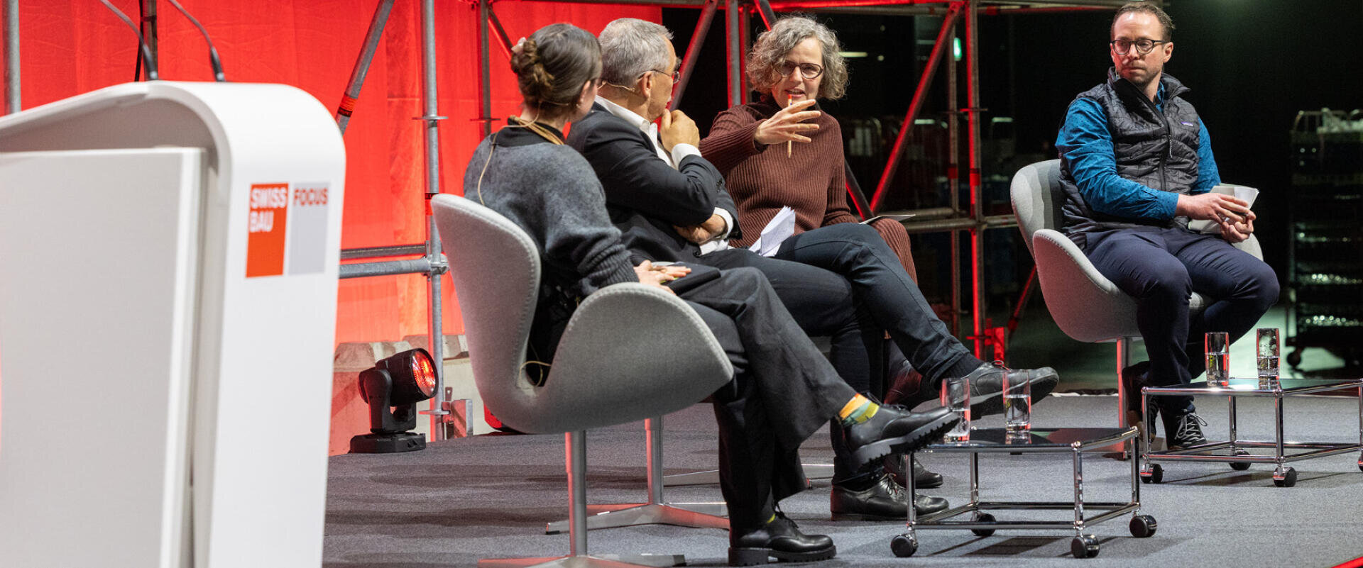 Vier Personen sitzen auf Stühlen auf einer Bühne und führen eine Podiumsdiskussion, wobei eine Frau gestikuliert, während sie spricht. Der Hintergrund ist rot, auf dem Podium prangt ein Schweizer Focus Bau-Schild. Es herrscht eine professionelle und aufmerksame Atmosphäre.